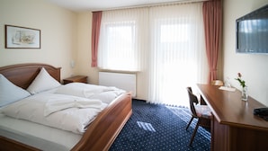 Una habitación de hotel con cama, escritorio, silla, radiador y ventana con cortinas.