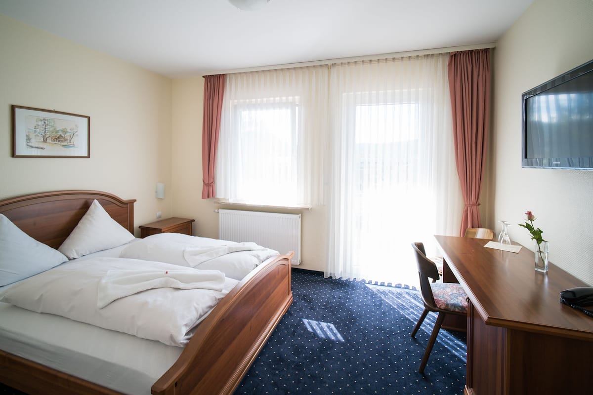 A hotel room with a bed, a desk, a chair, a radiator, and a window with curtains.