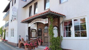 Un hotel con fachada europea tradicional, que incluye una señal con el nombre del hotel y restaurante, un arco de ladrillo y flores en macetas en la entrada.