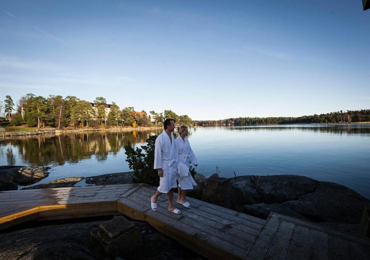 A couple in white robes walking on a wooden dock by a calm lake with trees and buildings in the background.