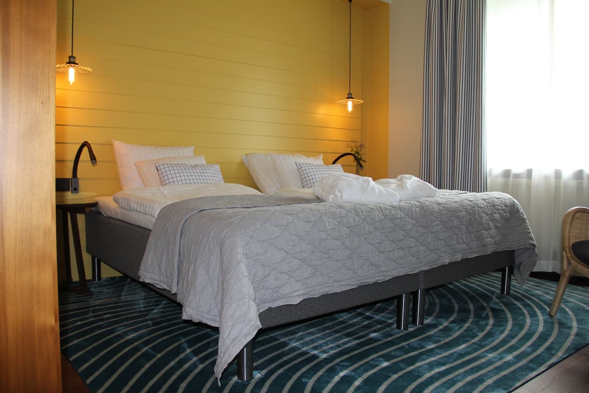 A double bed with a grey quilt, a bedside table with a lamp, and a window with curtains.
