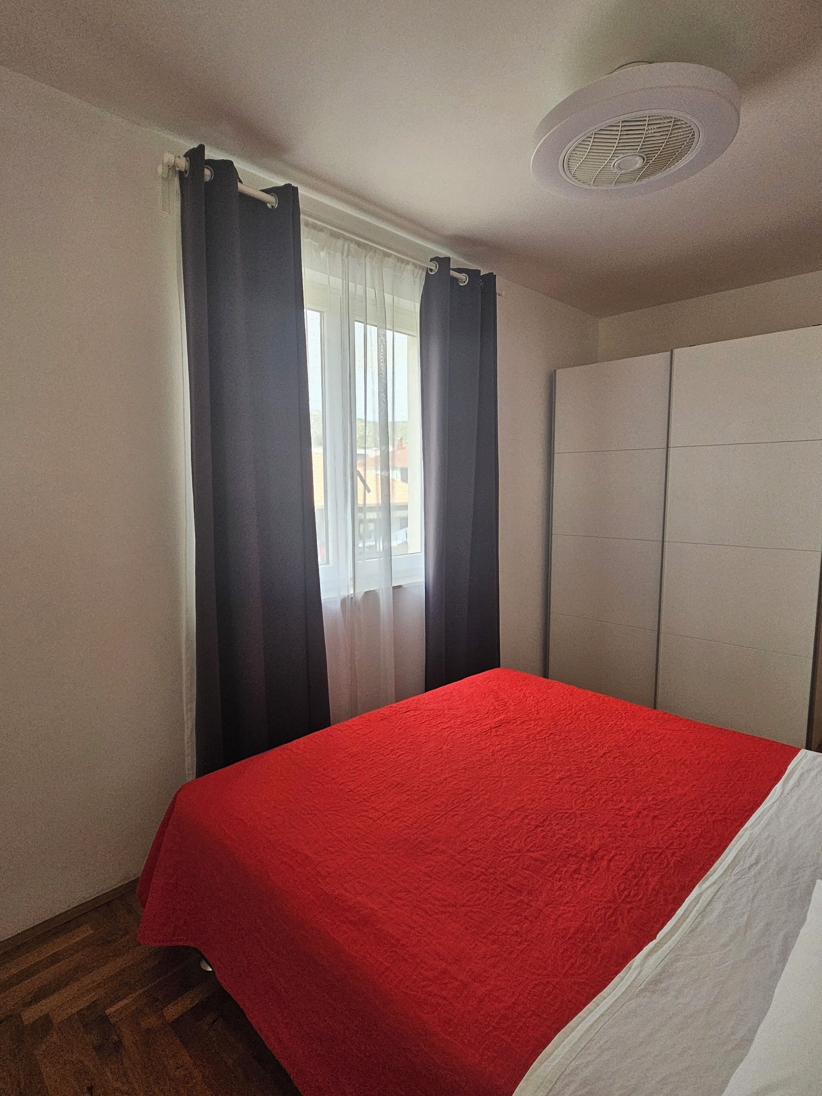 A room with a bed, a window with curtains, a ceiling vent, and a wardrobe.