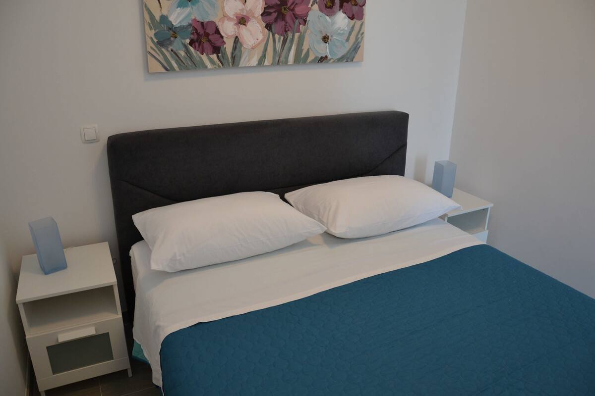 A neatly made bed with white pillows and a blue bedspread, a small bedside table with a blue object on it, and a wall-mounted painting of flowers.