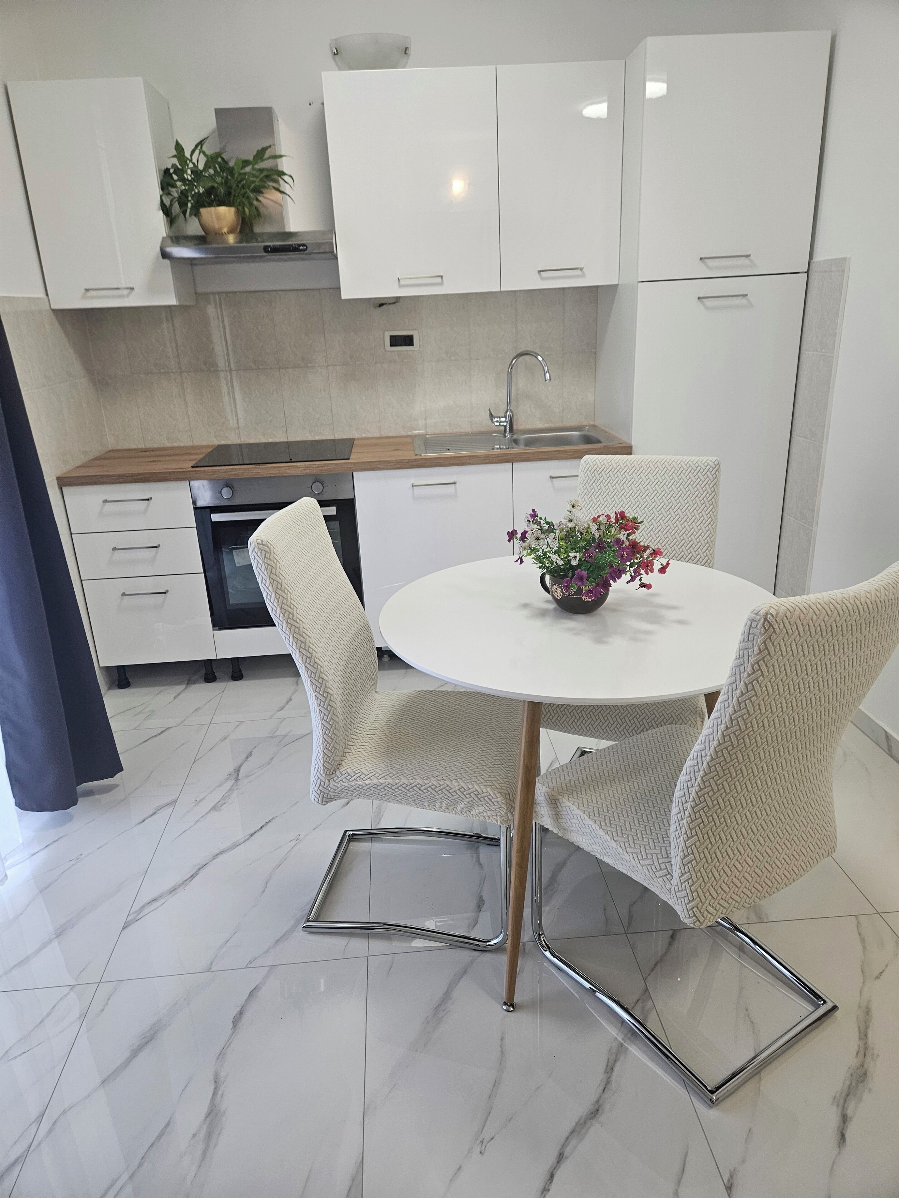 A modern kitchen with white cabinets, a stainless steel sink, and a built-in oven. There is a round dining table with four chairs and a vase of flowers.