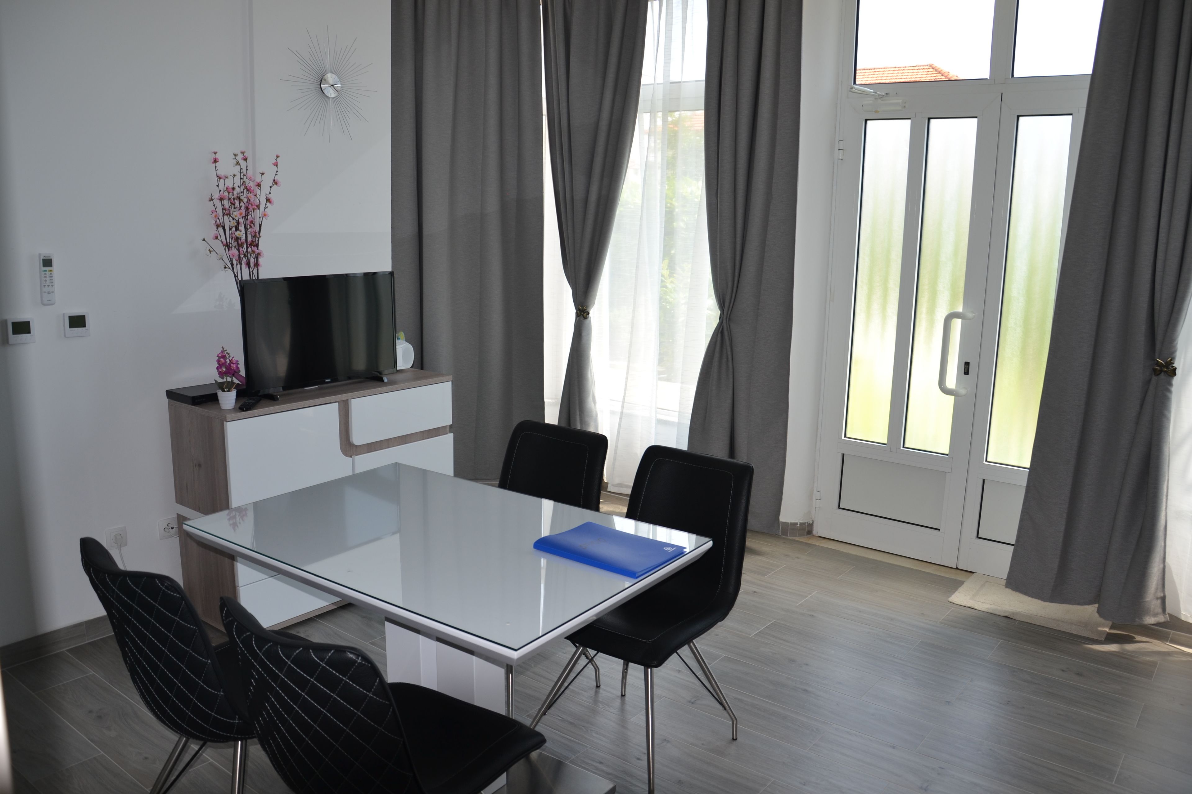 A modern dining area with a glass table, black chairs, a TV on a wooden cabinet, and a view through large windows with curtains.
