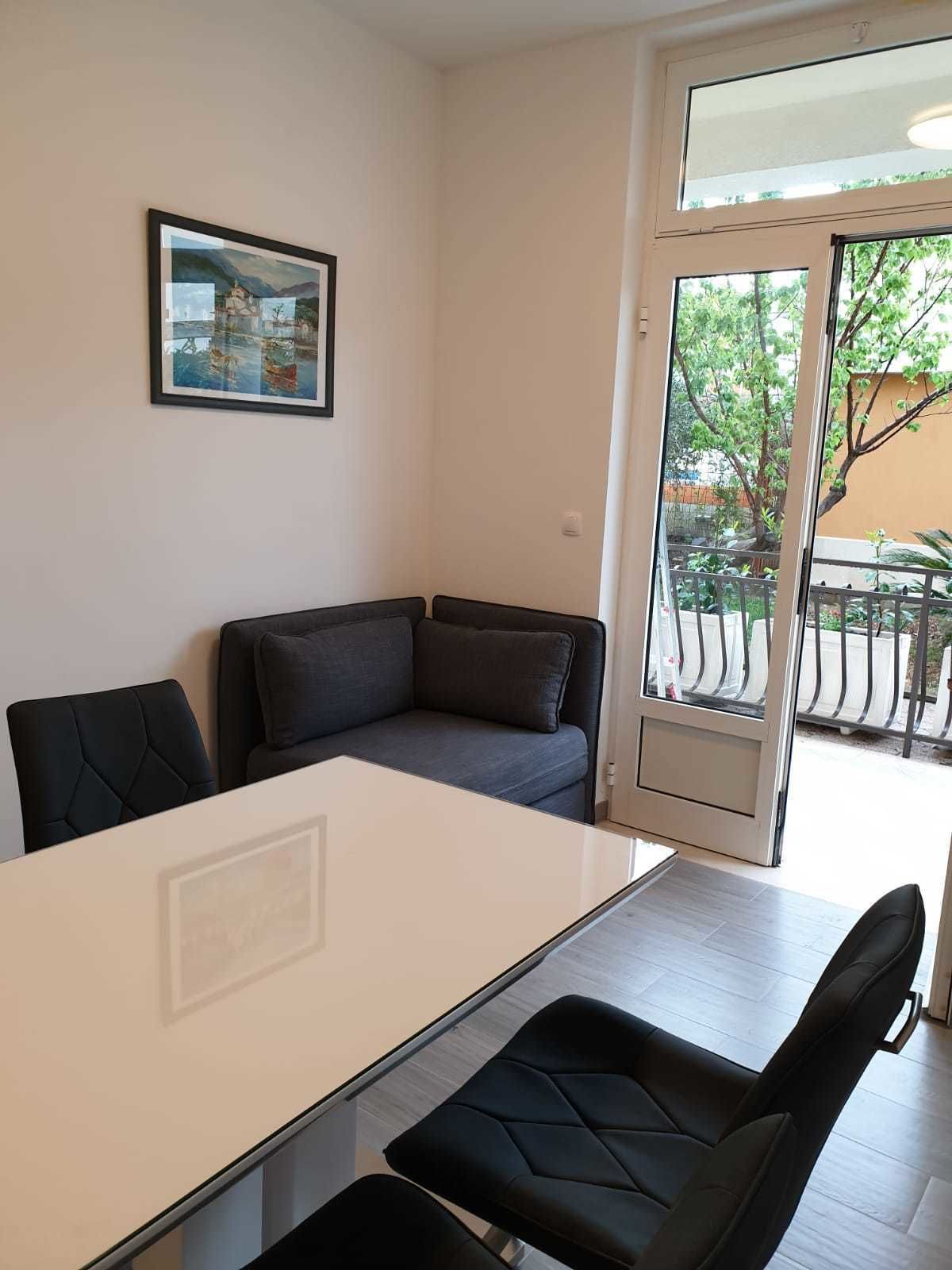 Una camera moderna con divano, tavolo da pranzo e sedie. Una porta scorrevole in vetro dà accesso a una zona esterna con vista su alberi e un edificio.