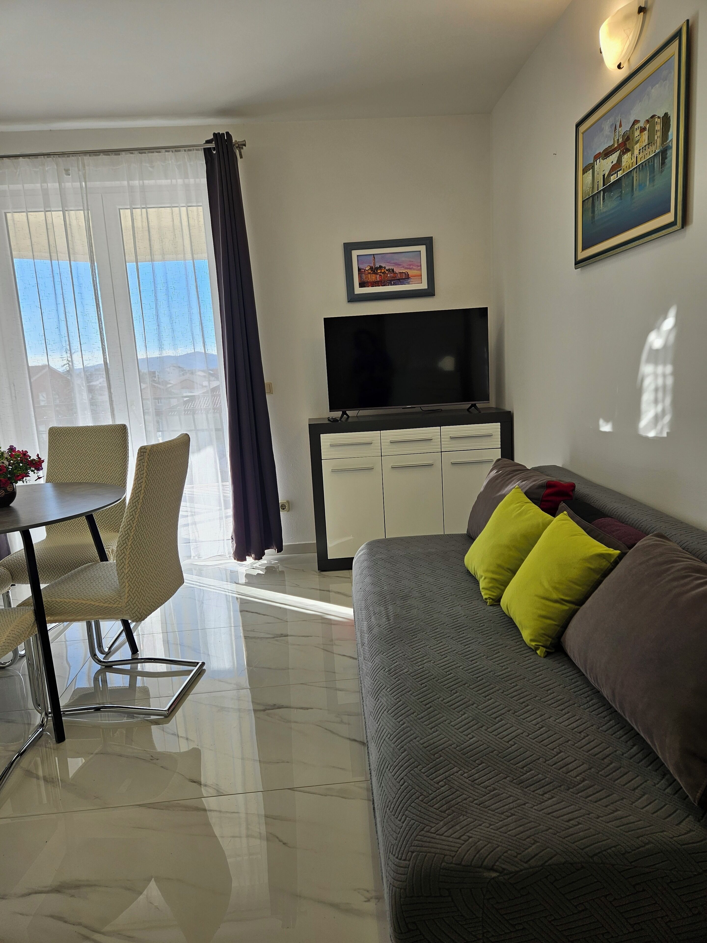 A modern living room with a sofa, a flat-screen TV, a dining table with chairs, and a large window with curtains.