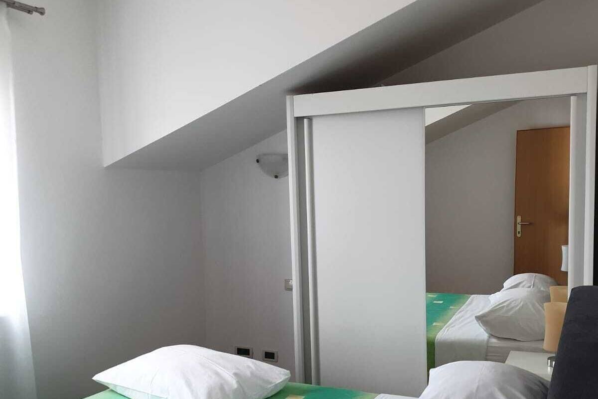 A small, single-bed room with a green and white bedspread, white pillows, a white bedside table, and a mirror on the wall.