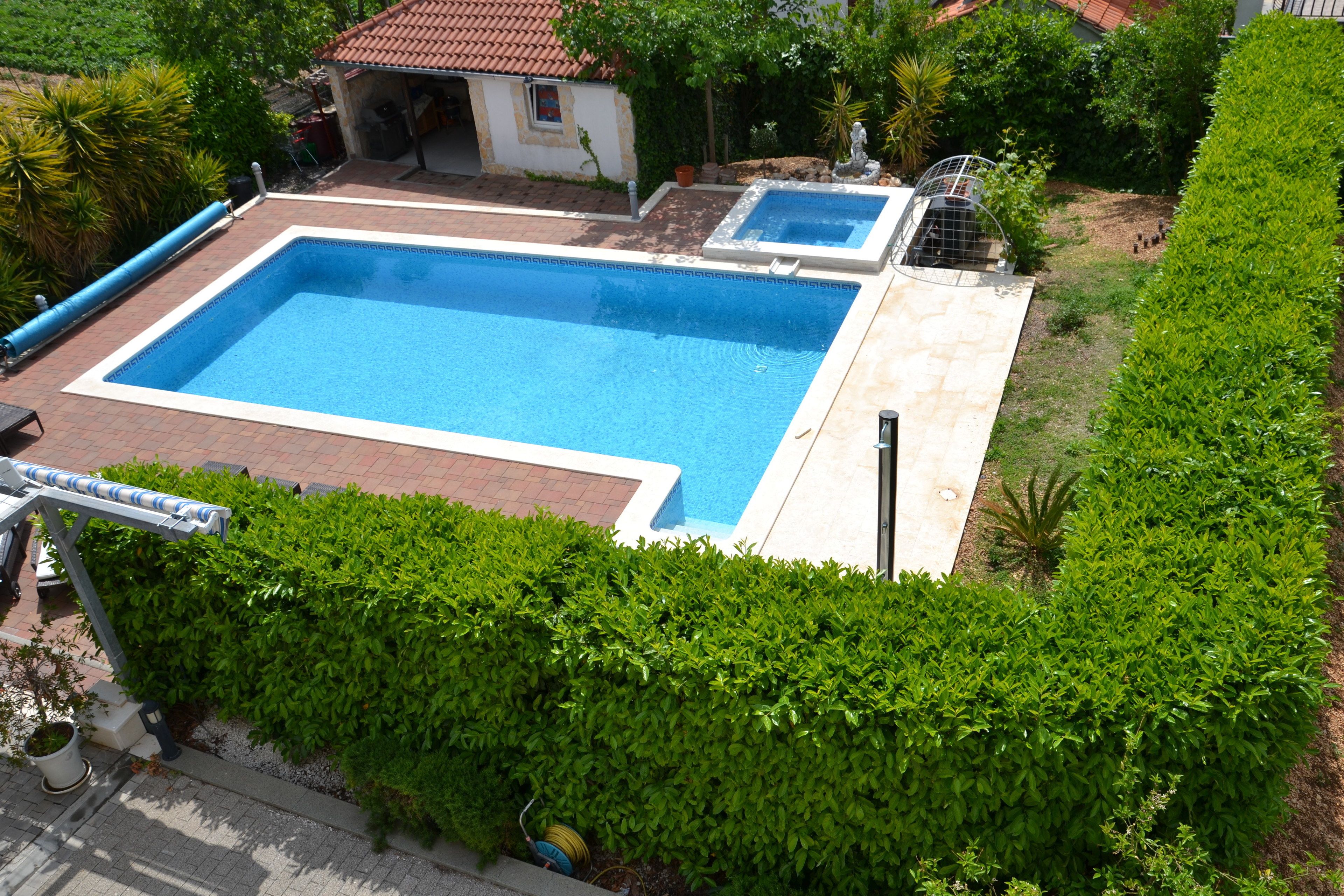 A rectangular swimming pool with a tiled deck, surrounded by well-maintained hedges and a small garden area.