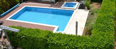 A rectangular swimming pool with a tiled deck, surrounded by well-maintained hedges and a small garden area.