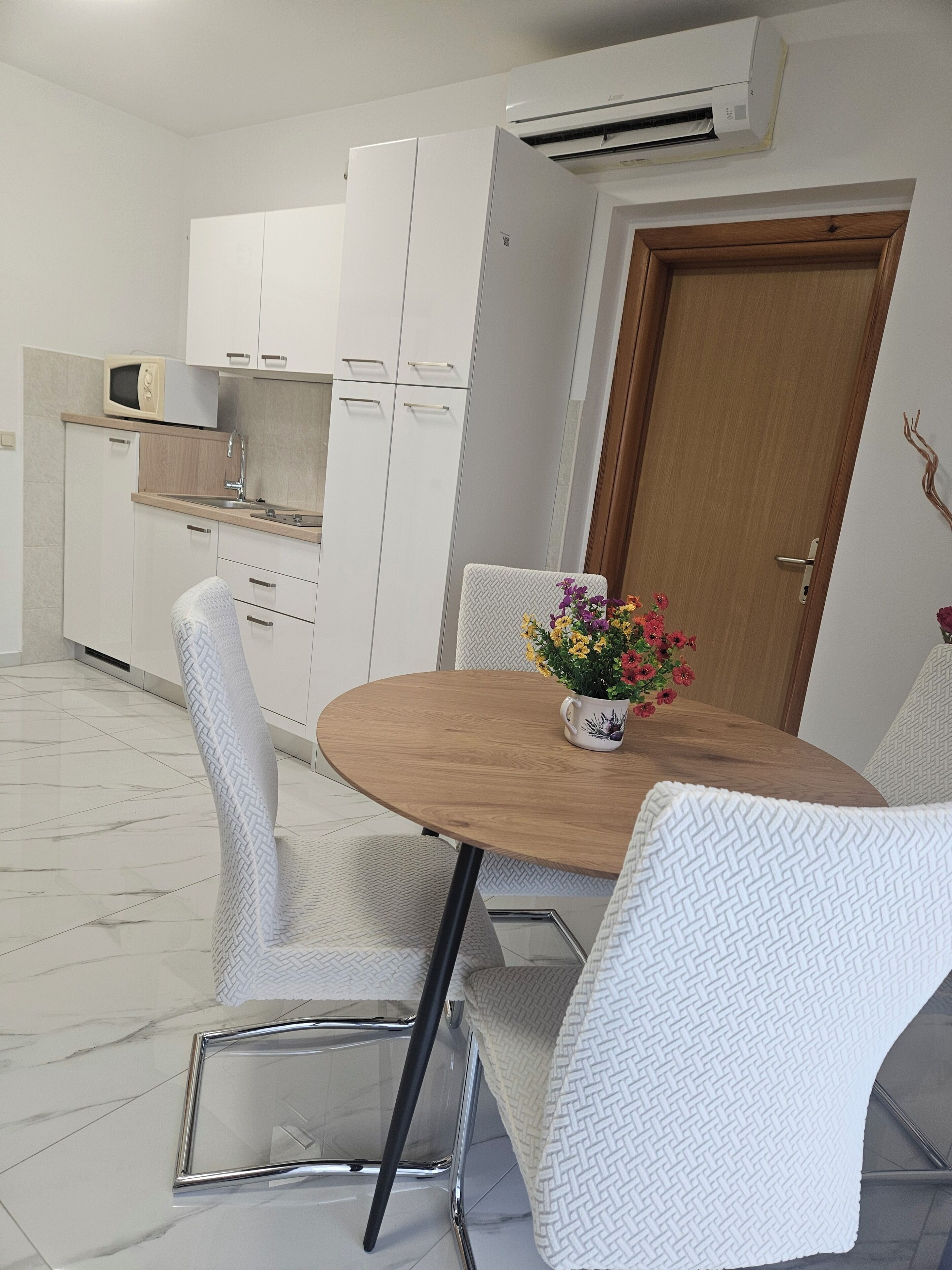 A modern kitchen with white cabinets, a microwave, and a sink. There is a wooden dining table with chairs and a vase of flowers.