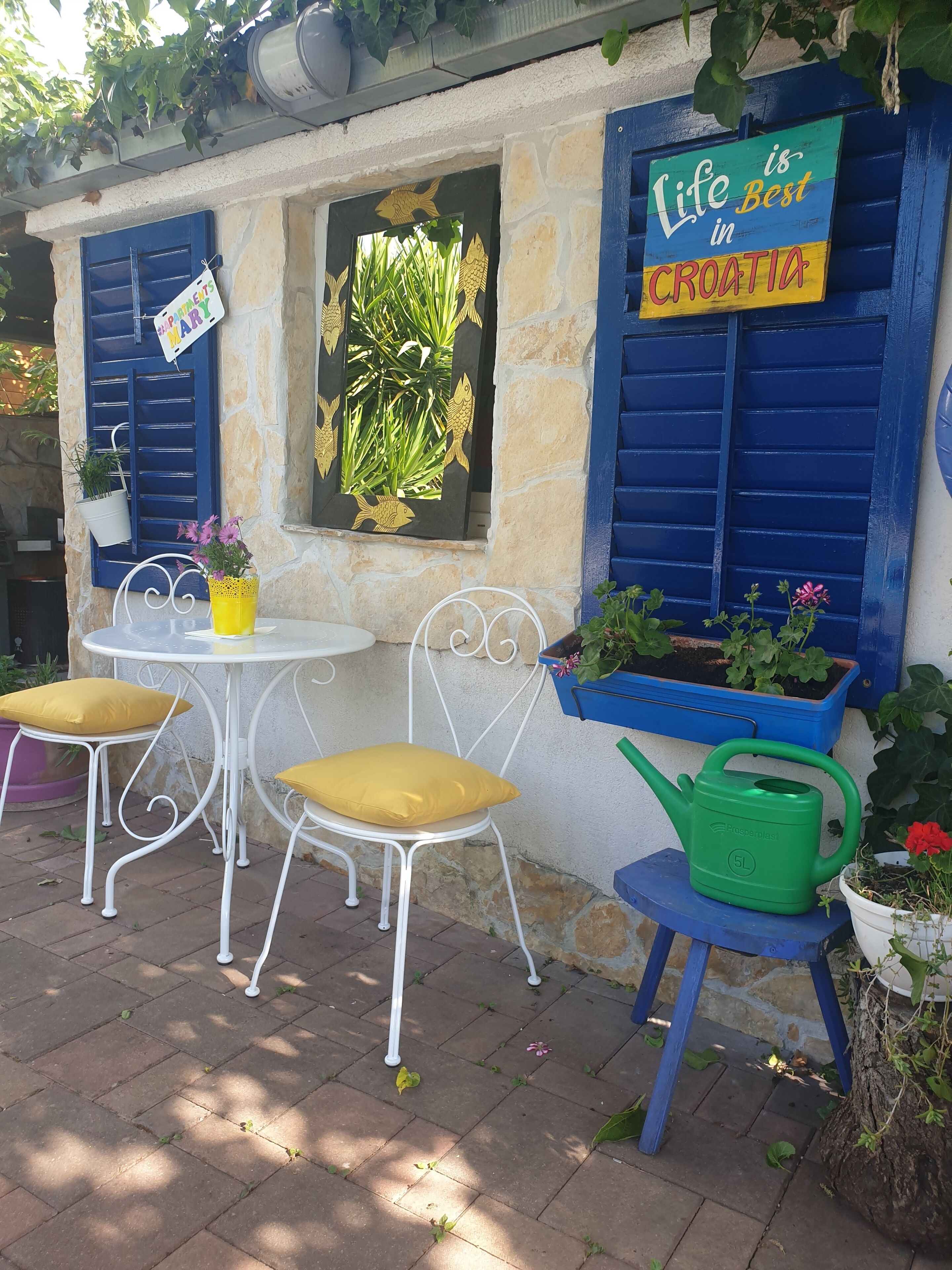 Un patio con pared de piedra, contraventanas azules, una mesa con cojines amarillos y un letrero que dice "Life is Best in Croatia".
