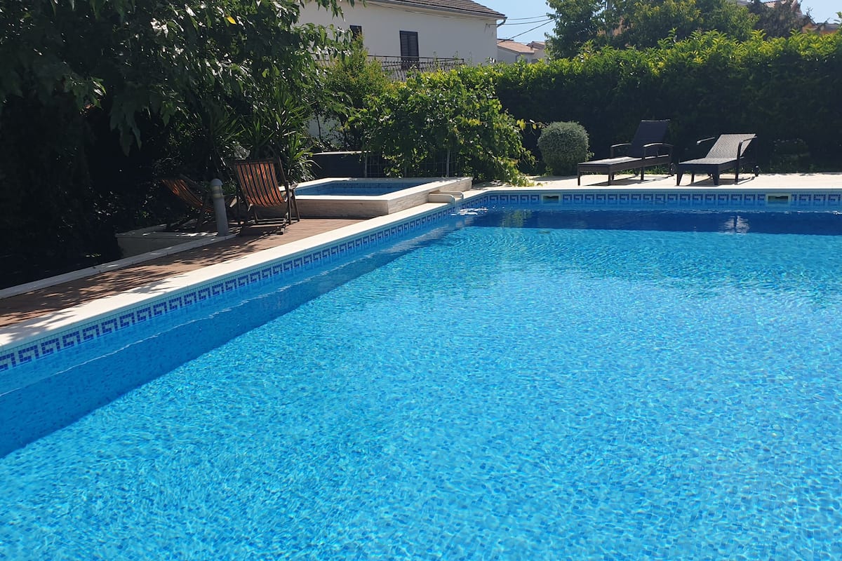 A clear blue swimming pool with a tiled edge, surrounded by lounge chairs and greenery.