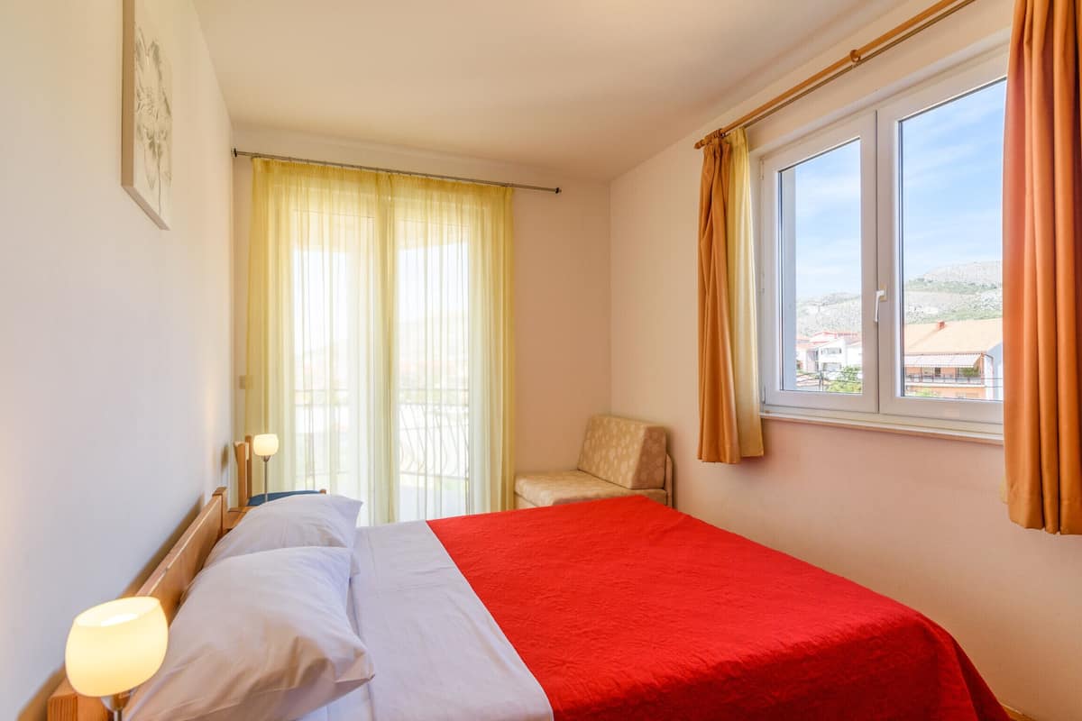 A hotel room with a bed, a chair, a window with curtains, and a lamp.