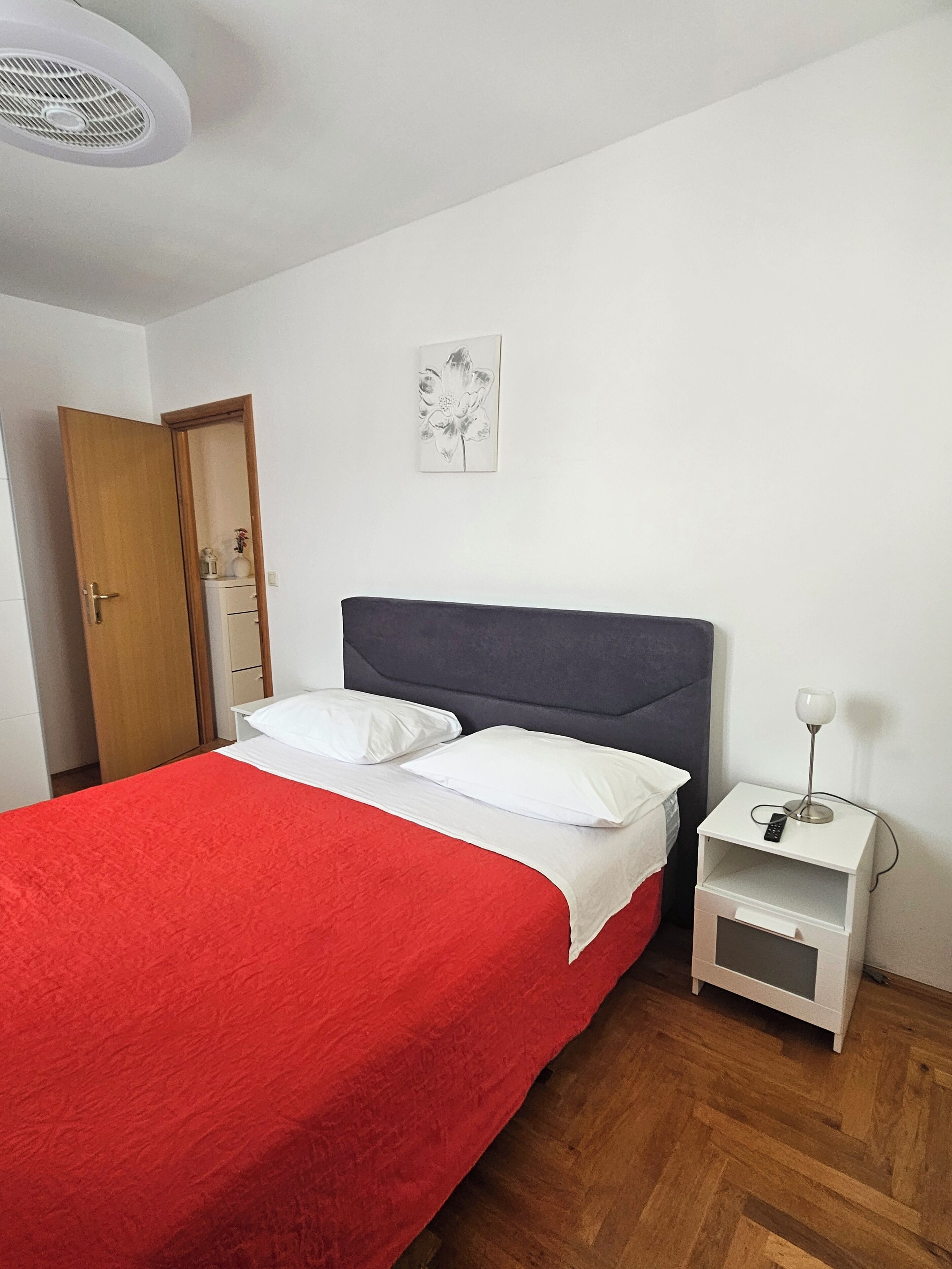 A bedroom with a bed, a nightstand, a wall-mounted lamp, a door, and a framed picture.