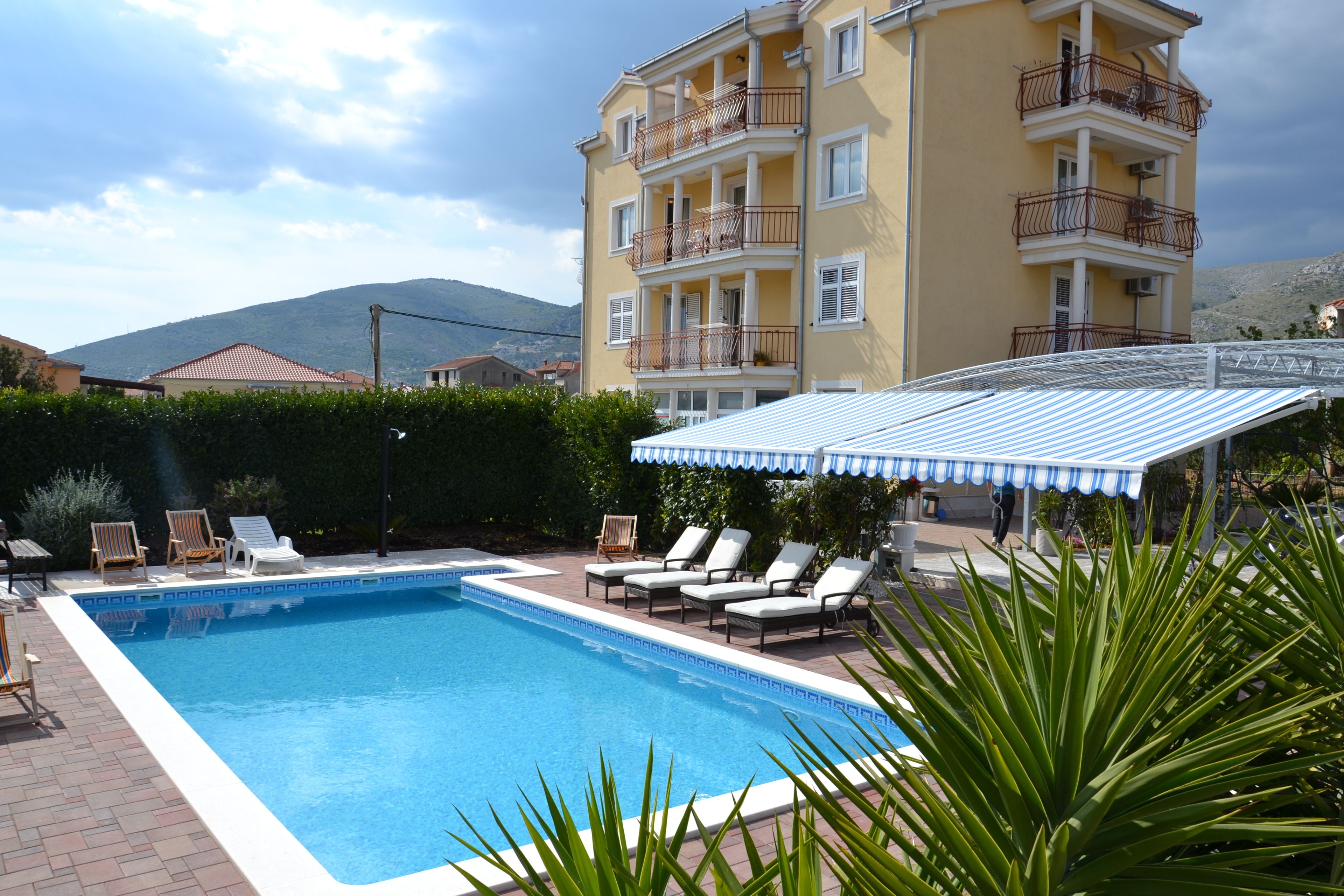 Una piscina con lettini e una zona coperta, circondata da un edificio con balconi e una montagna sullo sfondo.