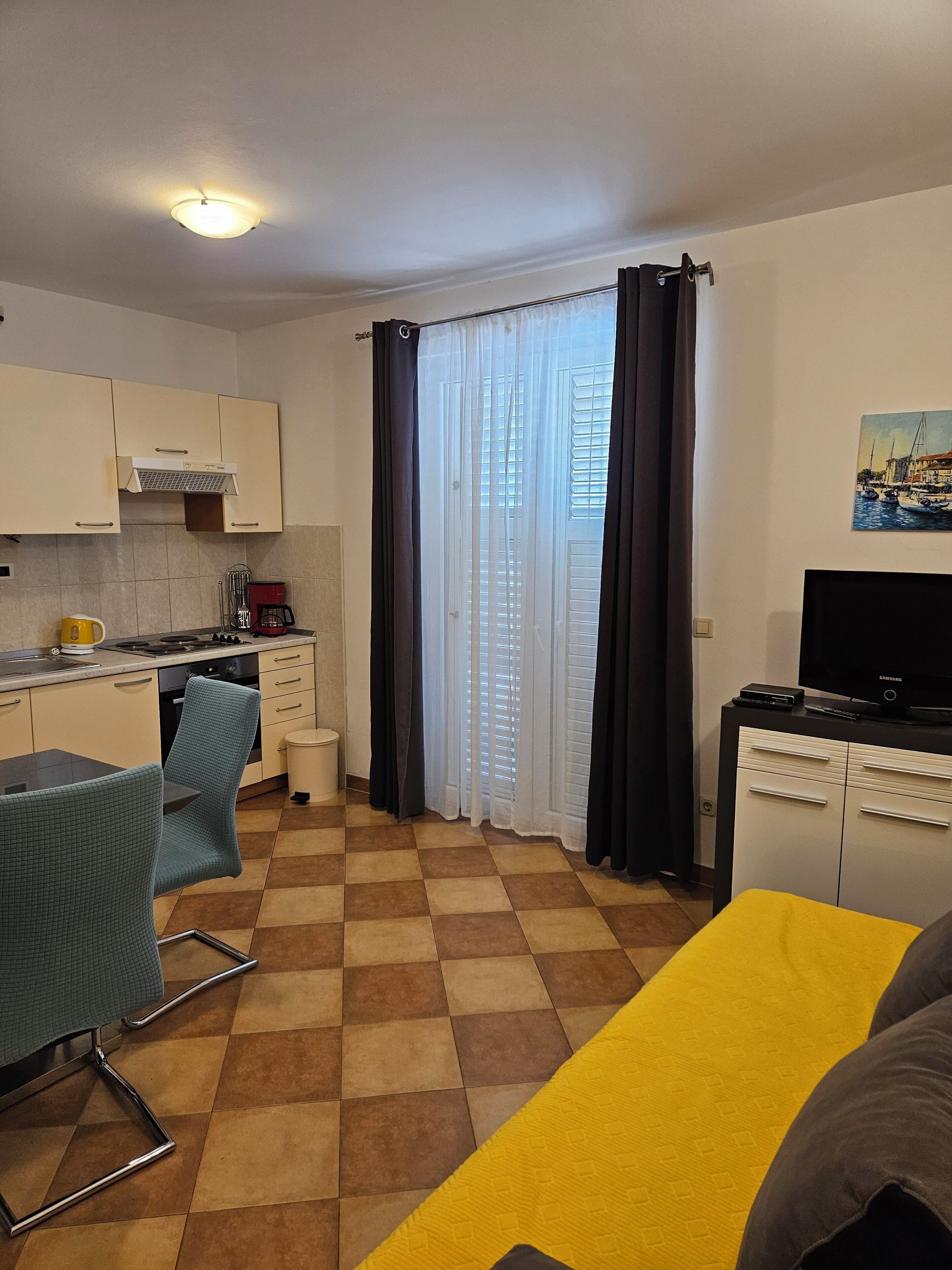 A compact kitchen with a stove, sink, and cabinets, a dining area with a table and chairs, a TV on a stand, and a window with curtains.