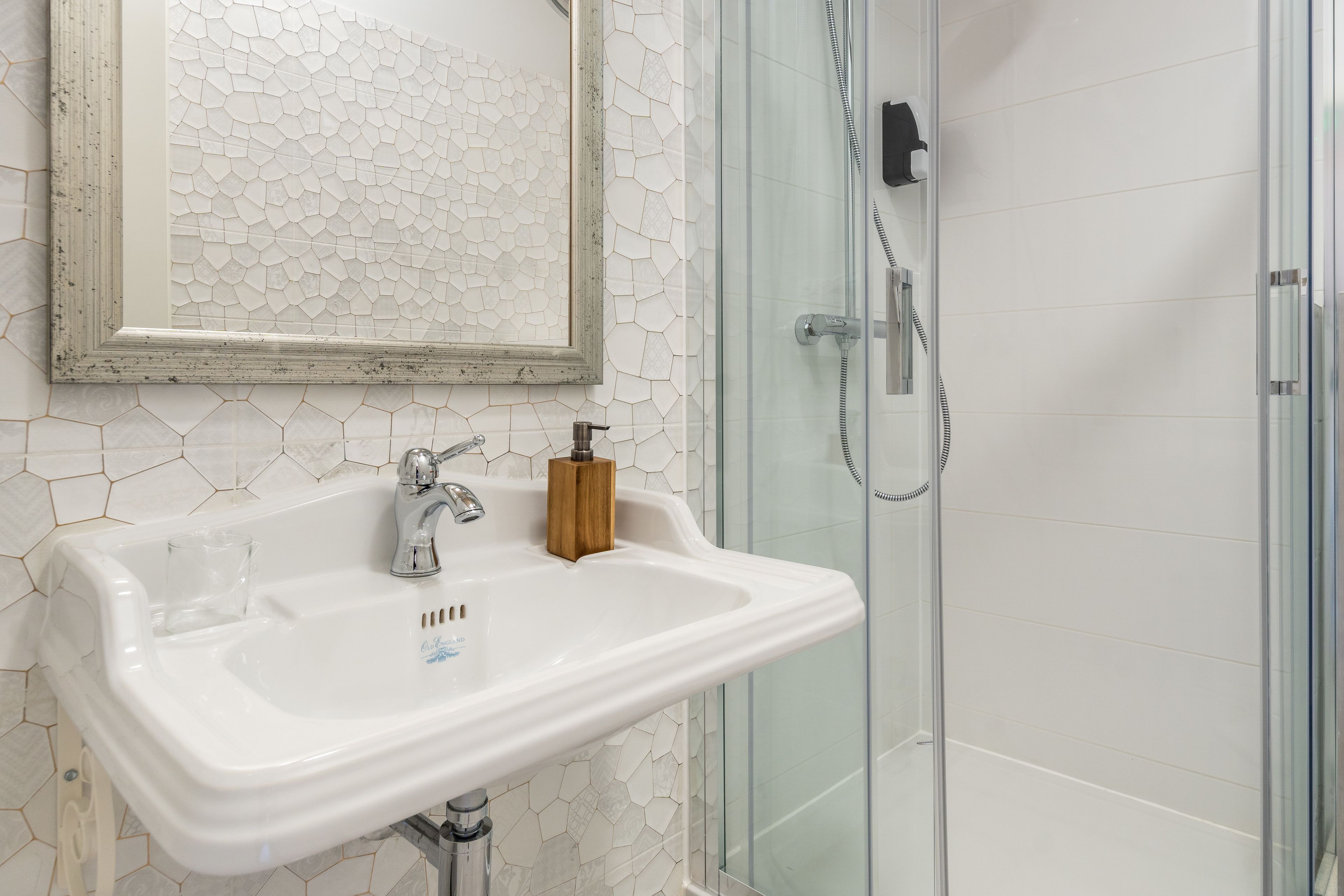 Een badkamer met een wit wastafel, een houten zeepdispenser, een douchecabine met glazen wanden en een spiegel met een achtergrond van hexagontegels.
