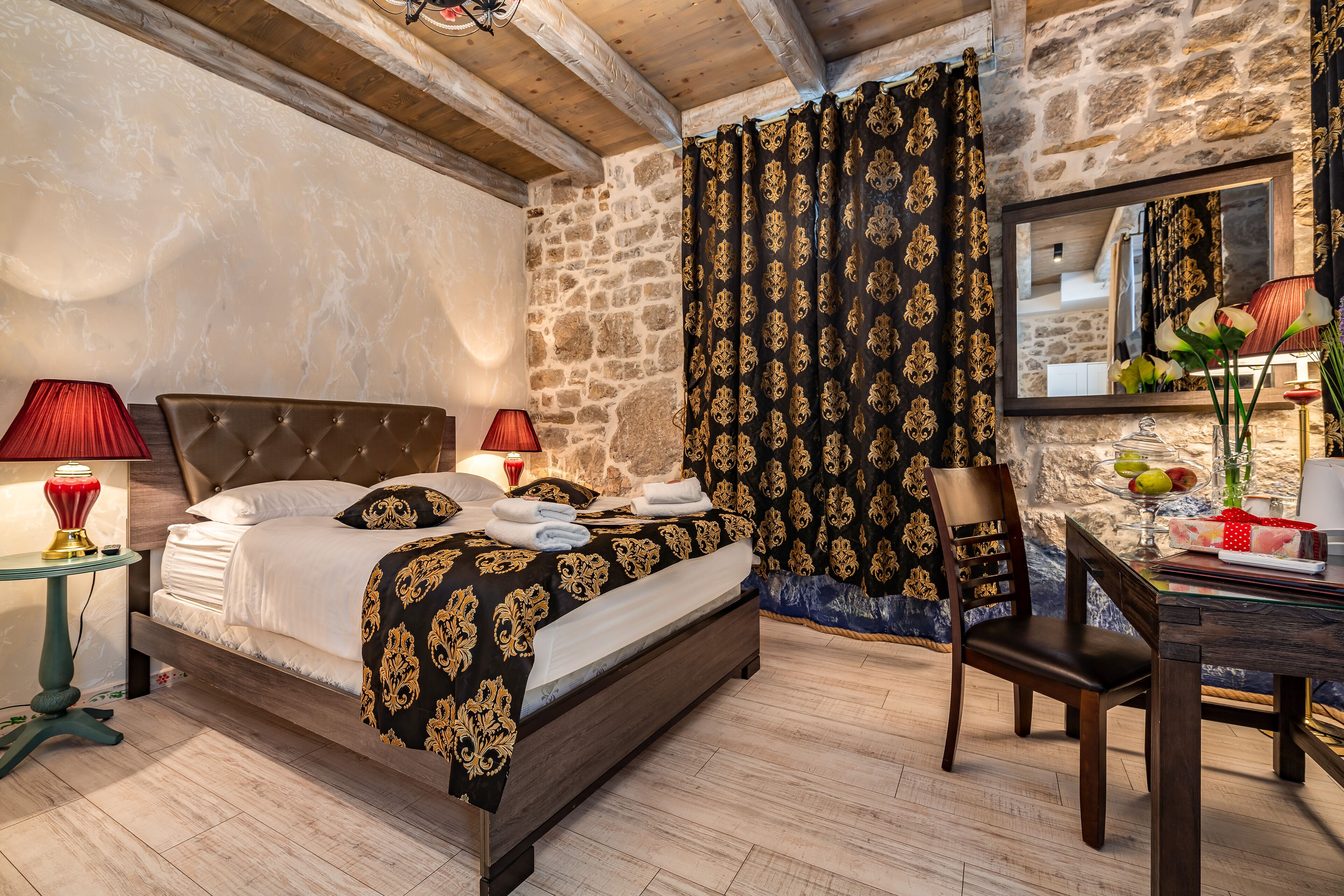 A bedroom with a bed, two bedside lamps, a chair, a desk with a mirror, and a stone wall.