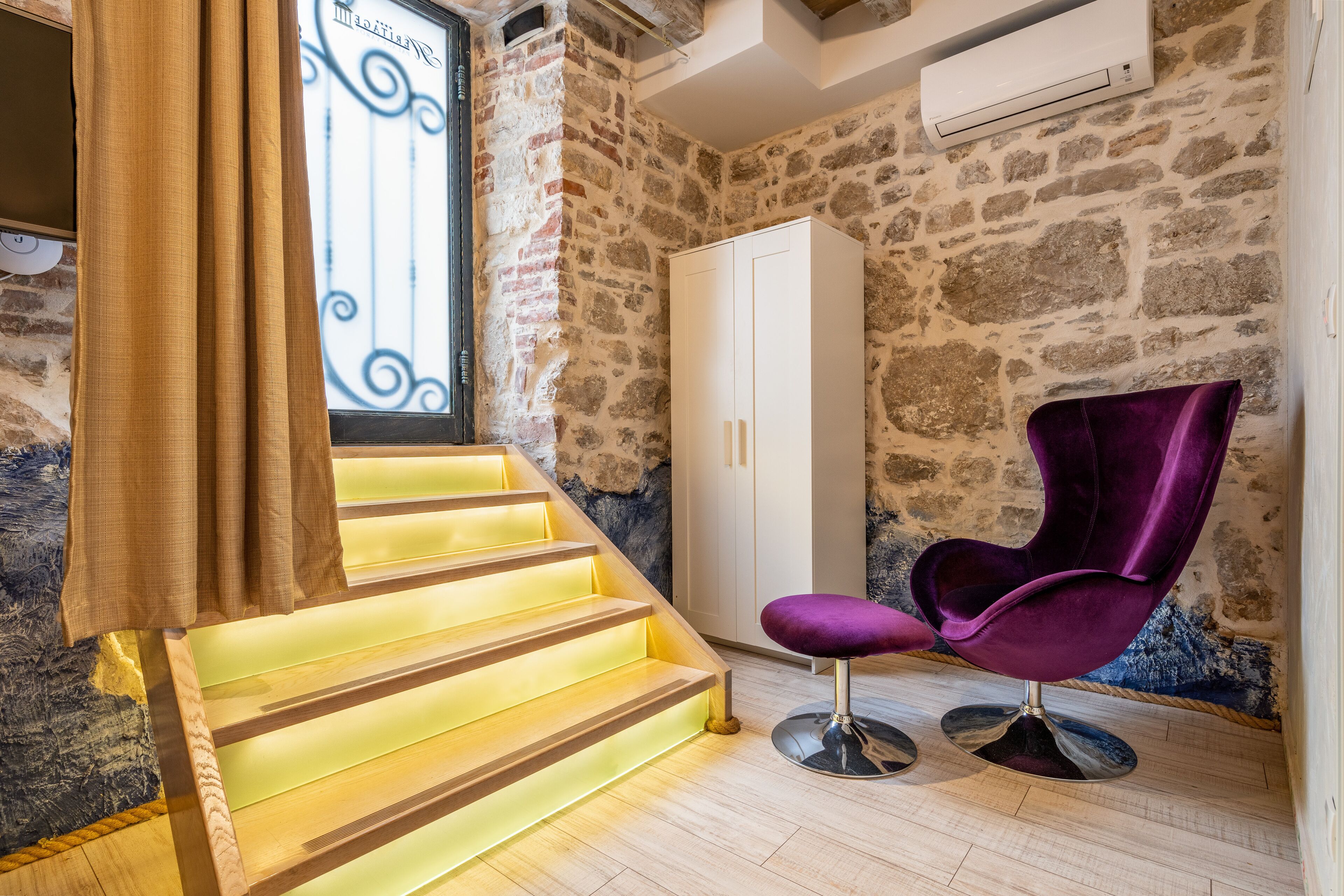 A staircase with illuminated steps, a white wardrobe, and a purple chair in a room with stone walls and a window with decorative ironwork.
