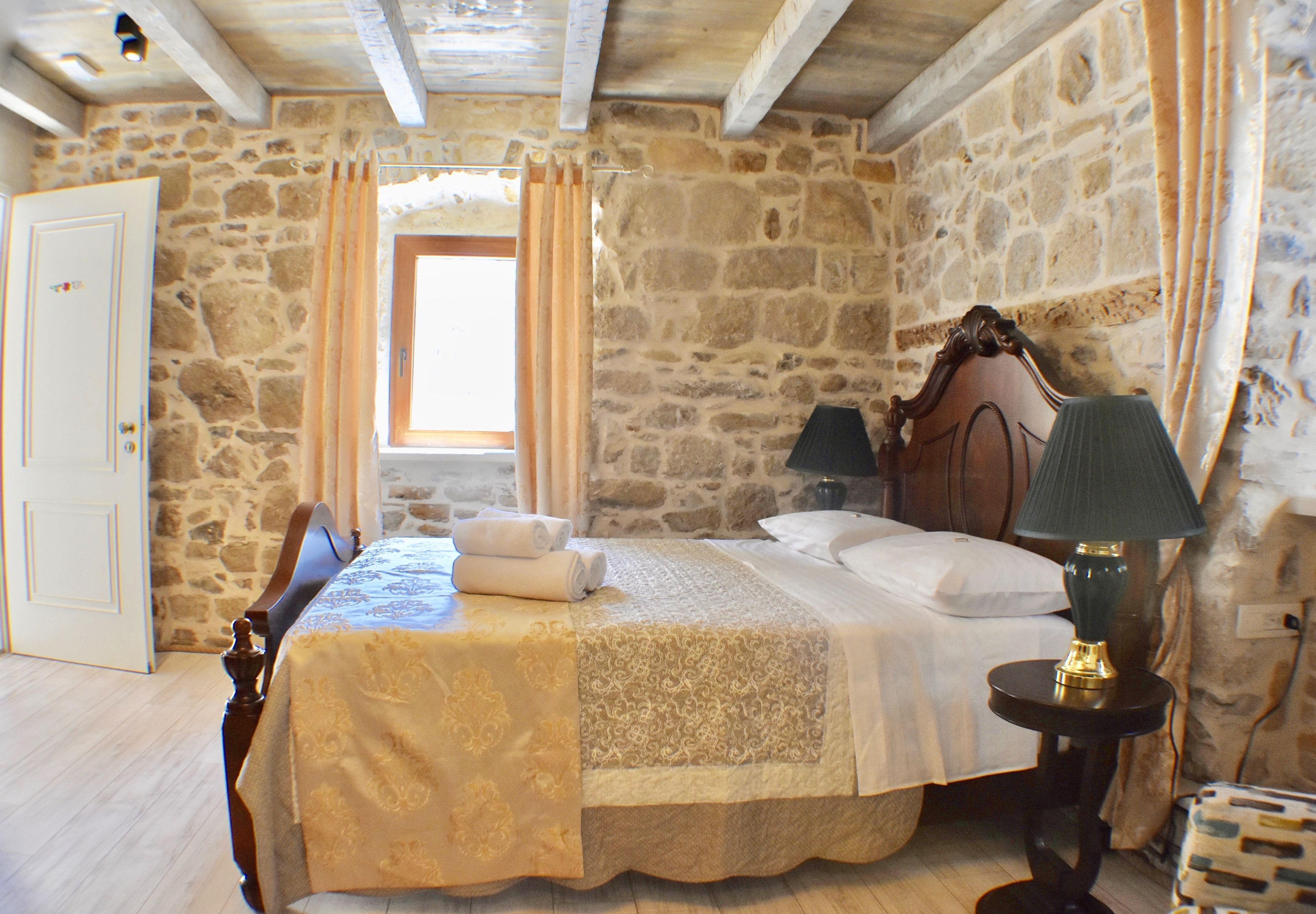 A bedroom with a stone wall, a wooden bed, a nightstand, a lamp, and a window with curtains.