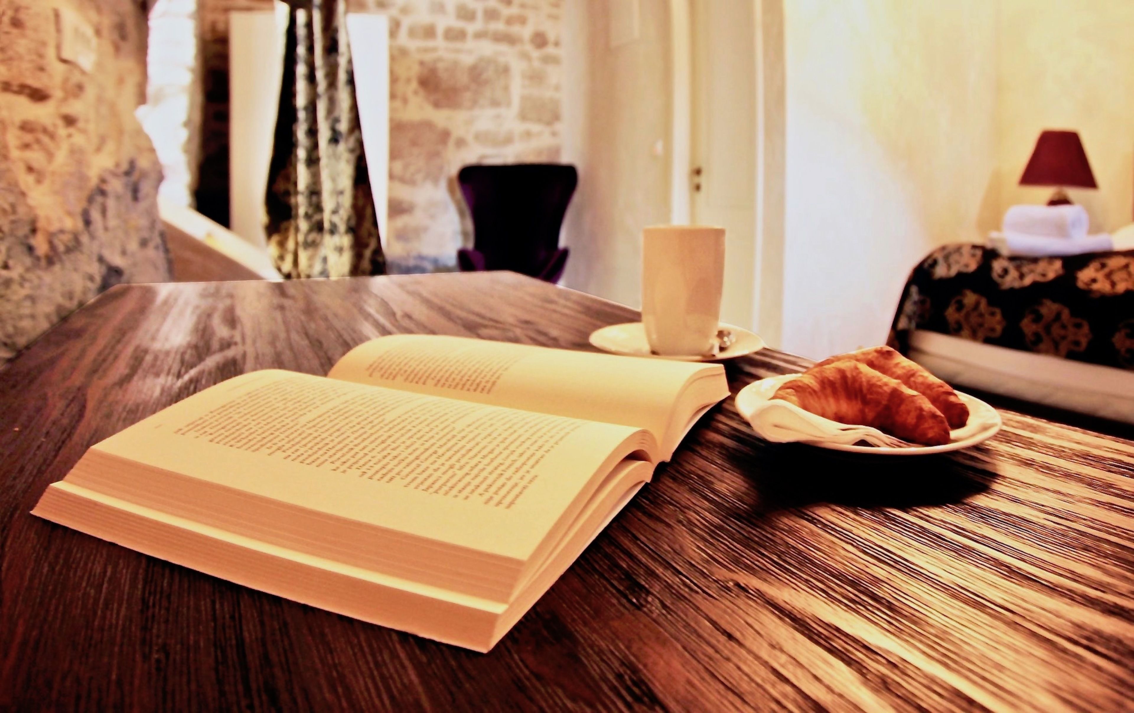 An open book on a wooden table with a croissant and a cup.