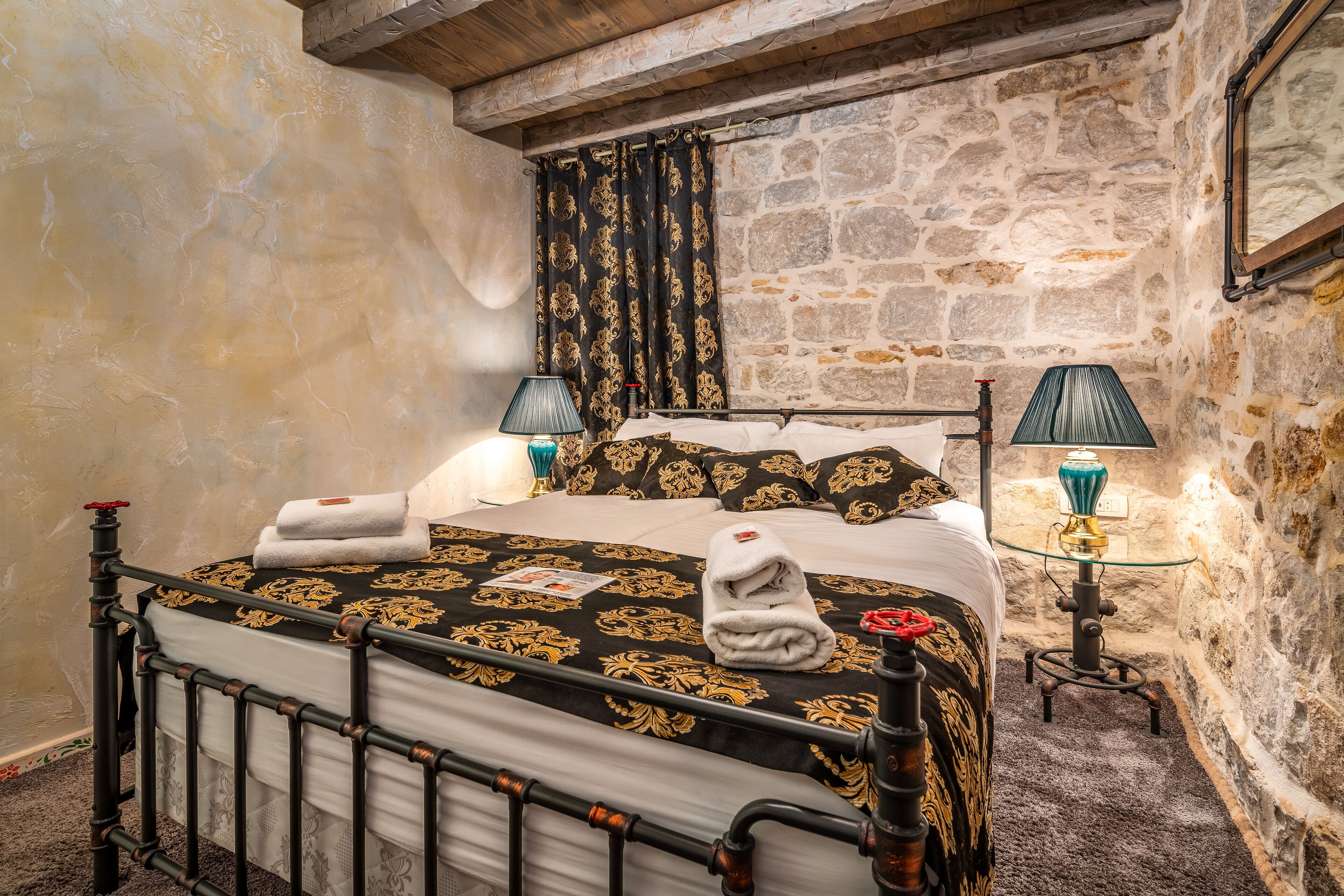 Een slaapkamer met een stenen muur, een bed met een zwart-gouden dekbedovertrek, twee bedlampen en een houten hoofdeinde.