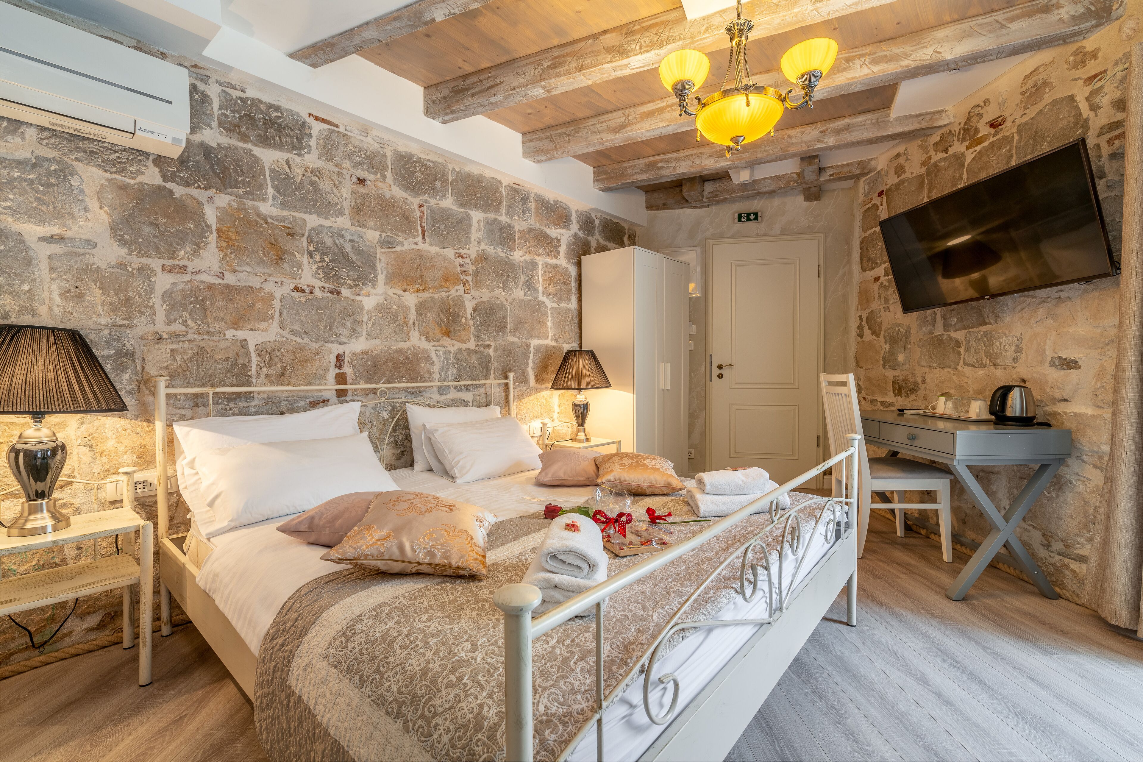 A bedroom with a stone wall, a bed with white bedding, a bedside table with a lamp, and a TV mounted on the wall.