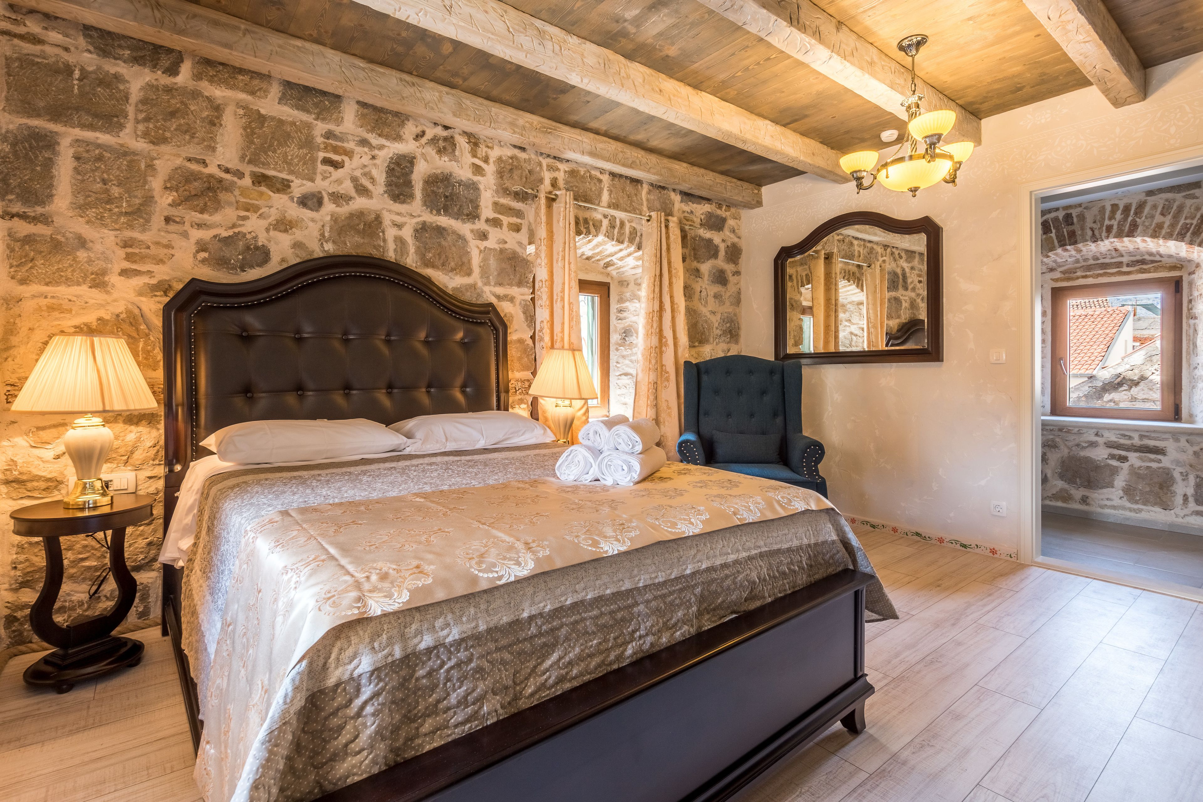 A bedroom with a stone wall, a wooden ceiling, a large bed with a headboard, a chair, a lamp, and a mirror.