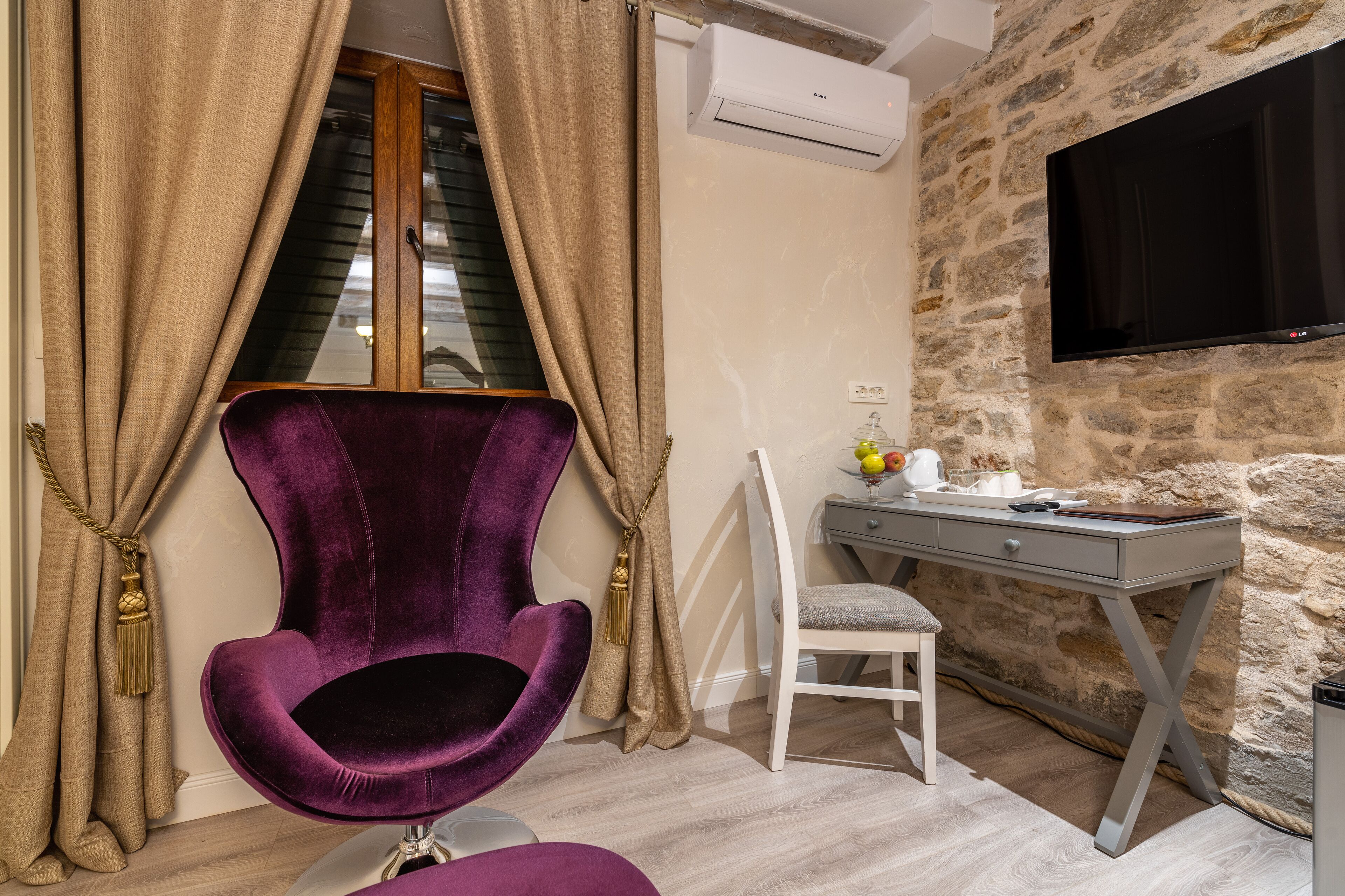 A room with a stone wall, a mounted TV, a desk with a chair, and a purple chair.
