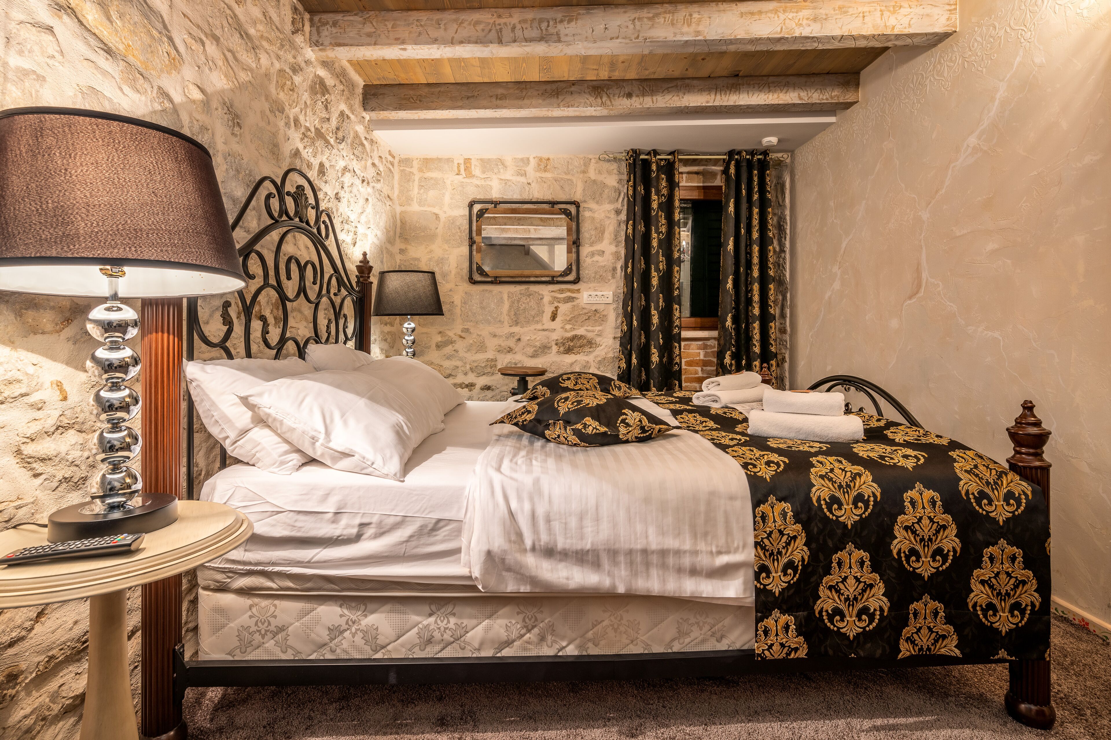 A bedroom with a stone wall, a bed with white and patterned bedding, a lamp, and a mirror.