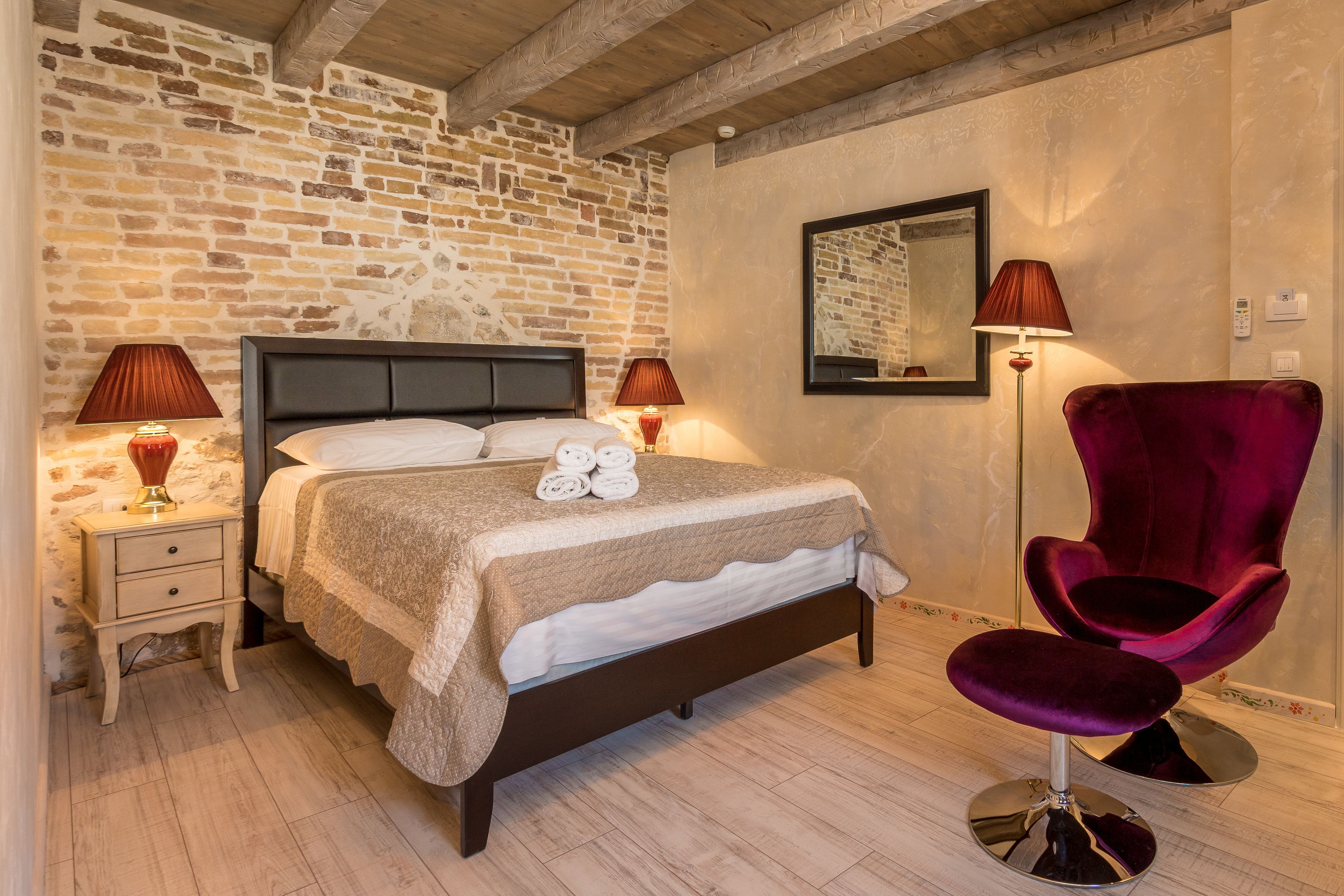 Un dormitorio con pared de ladrillo, una cama con cabecera, dos lámparas de noche, una silla roja y un espejo en la pared.