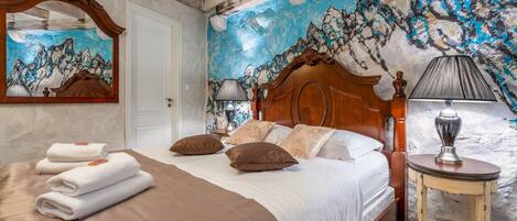 A bedroom with a bed, pillows, a bedside table with a lamp, a mirror, and a wall mural.