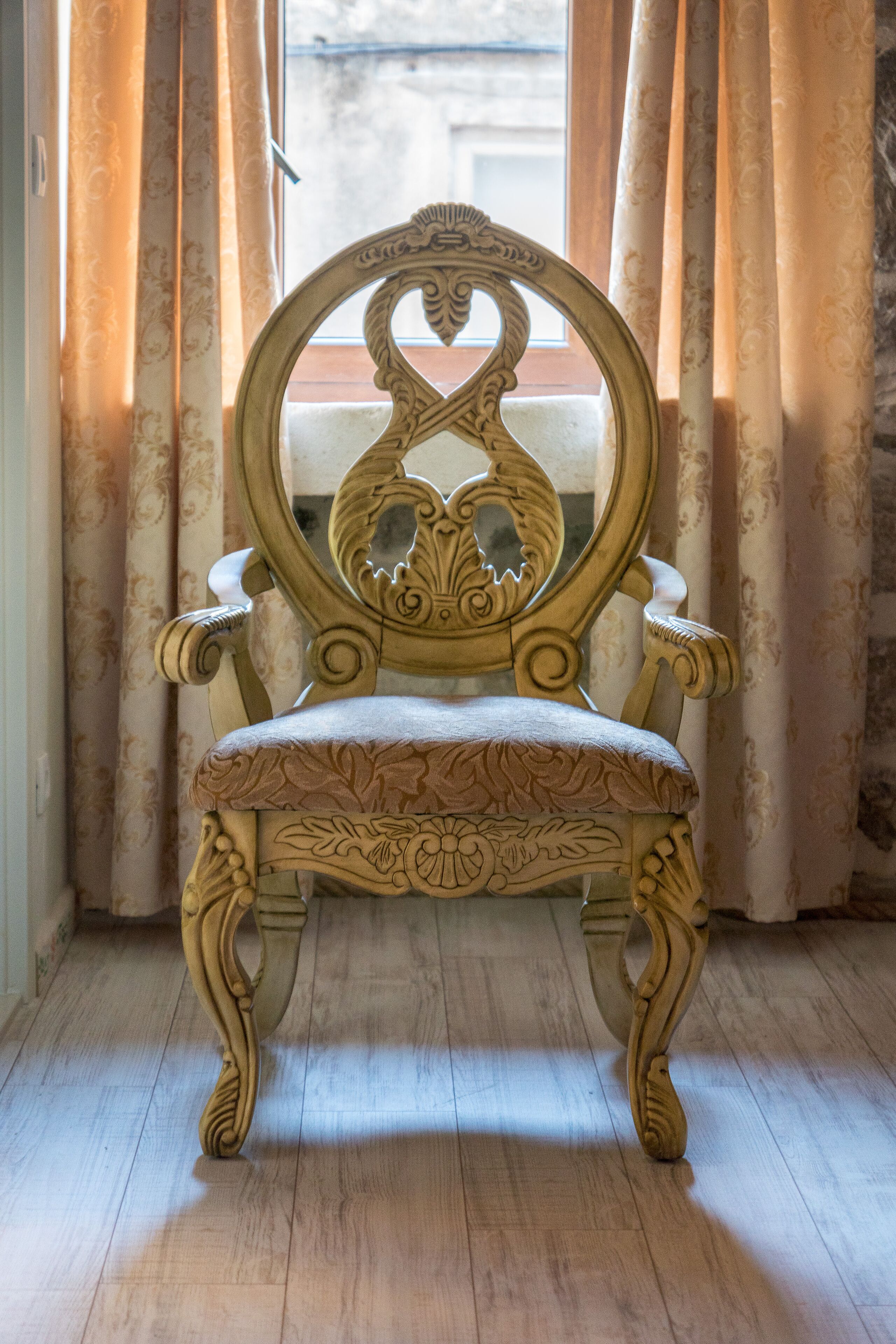 Una silla antigua con elaborados adornos, ubicada en una habitación con suelo de madera y cortinas.