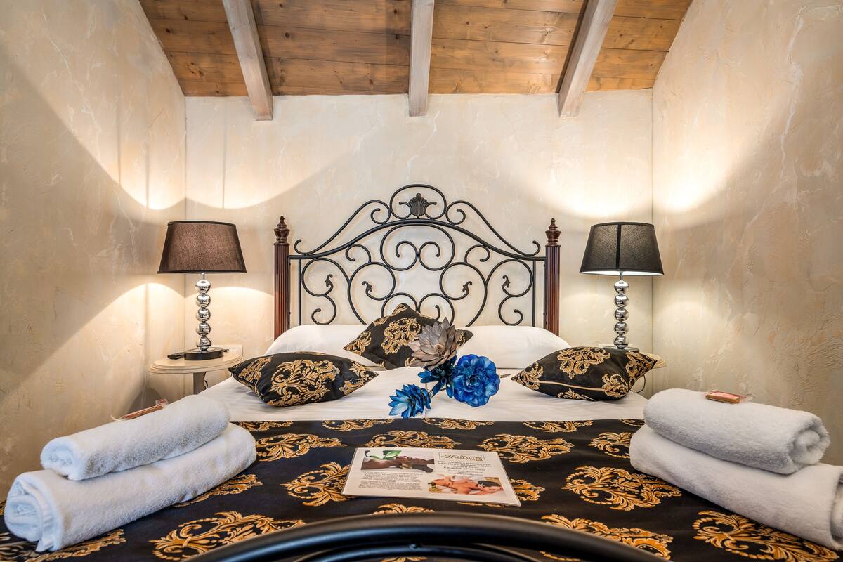 Ein ordentlich bezogenes Bett mit einer schwarz-goldenen Tagesdecke, zwei Kissen mit einem passenden Design und einer blauen Blumenanordnung. Zwei Nachttische mit schwarzen Lampenschirmen, und ordentlich gefaltete Handtücher liegen auf dem Bett.