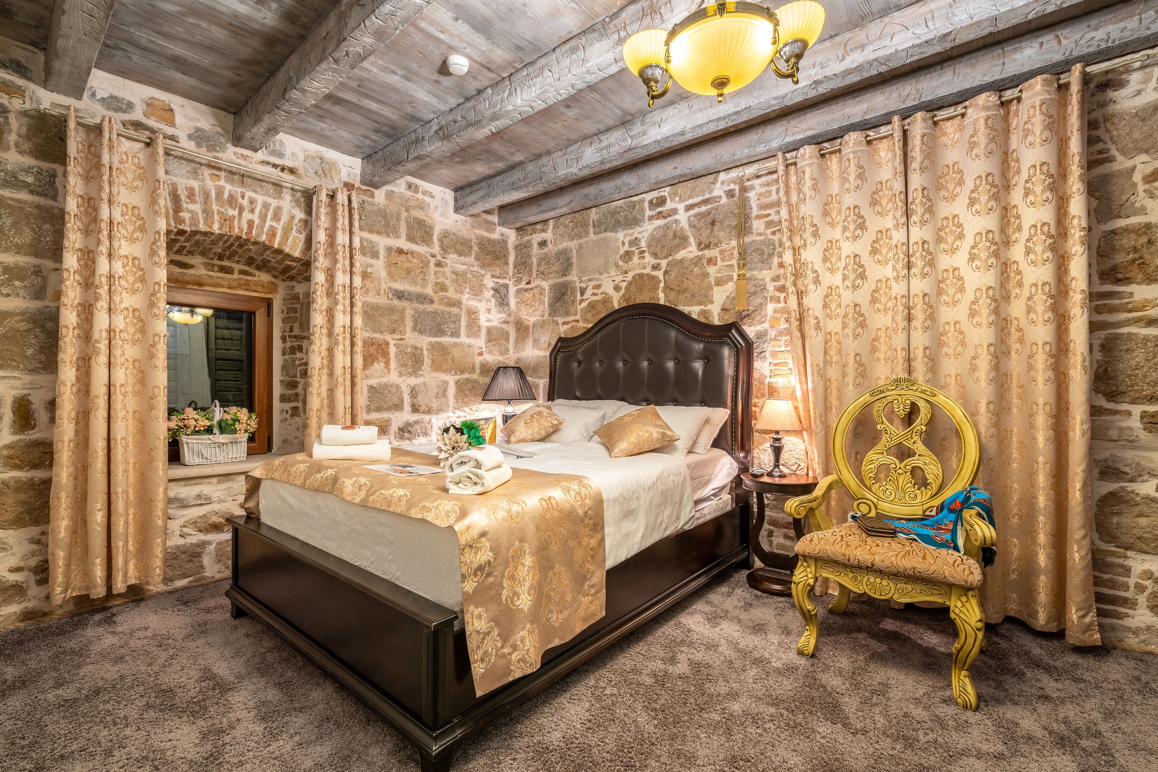 Une chambre aux murs de pierre, avec un grand lit, une tête de lit en bois, une table de chevet, une chaise et un lustre.