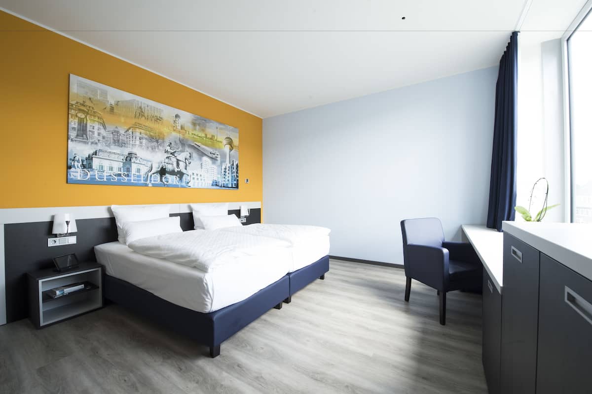 Habitació d'hotel moderna amb un llit gran, un escriptori i una pintura de paisatge urbà a la paret.