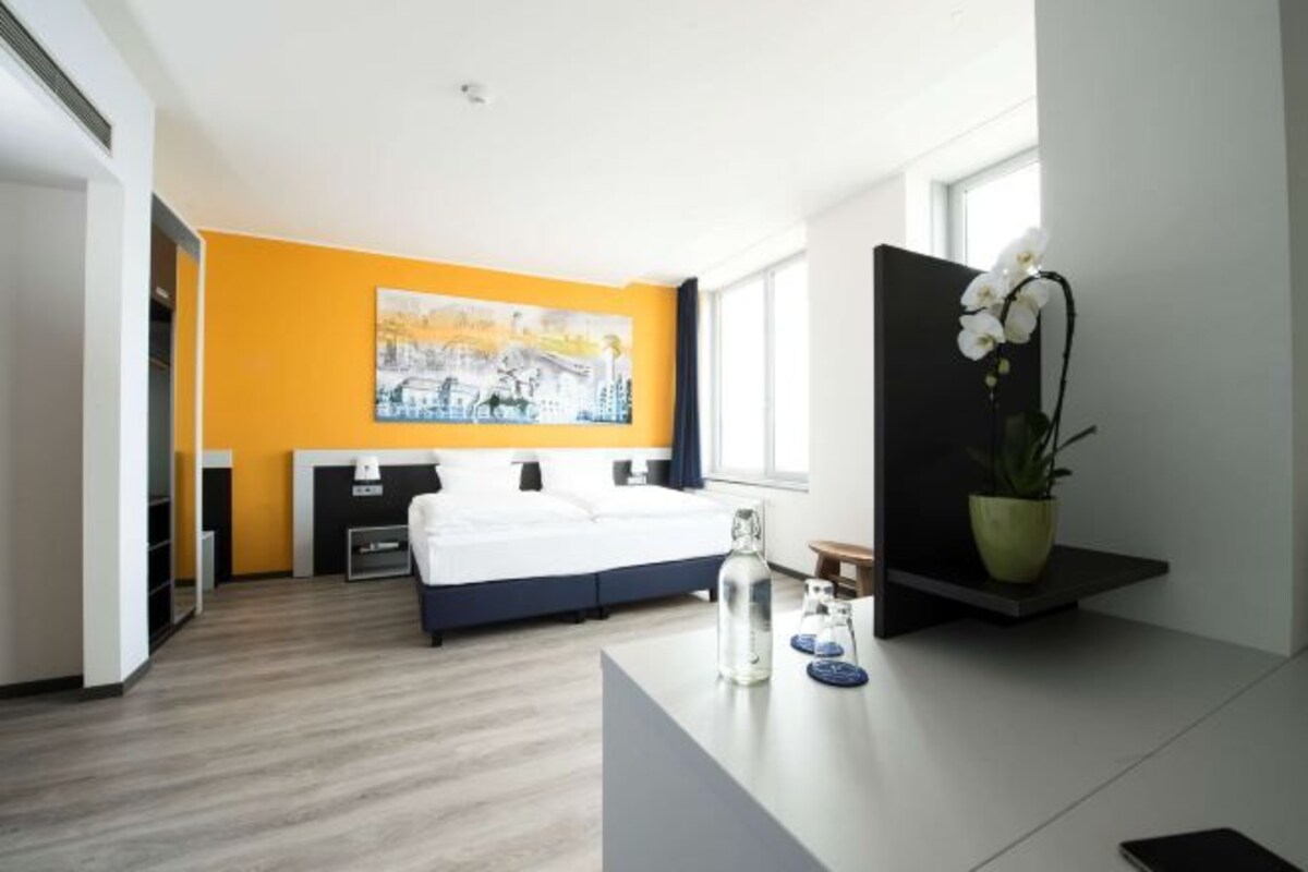 Ein modernes Hotelzimmer mit einem Bett, einem Bild an der Wand, einer Zimmerpflanze und einem Nachttisch mit einer Wasserflasche und Gläsern.
