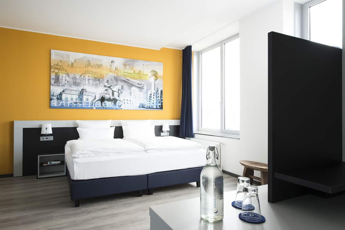 Una habitació d'hotel amb un llit, un quadre a la paret, un televisor, una taula de nit amb una ampolla i una finestra.