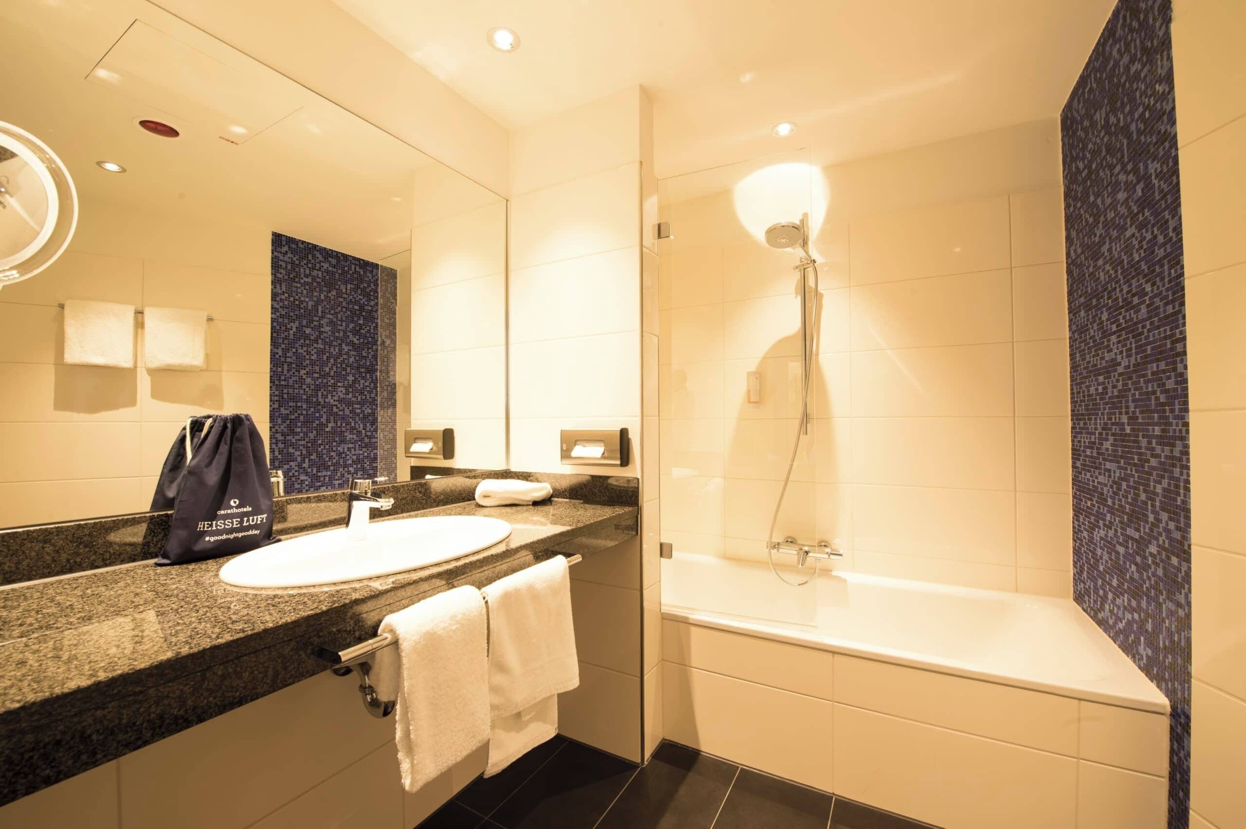 Un baño moderno con un lavabo redondo, un espejo, un perchero para toallas, una ducha con cabezal de ducha manual y una pared de mosaicos oscuros.