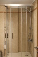 Cabina de ducha con paredes de vidrio, un cabezal de ducha de mano, un grifo empotrado en la pared y un dispensador de papel higiénico.