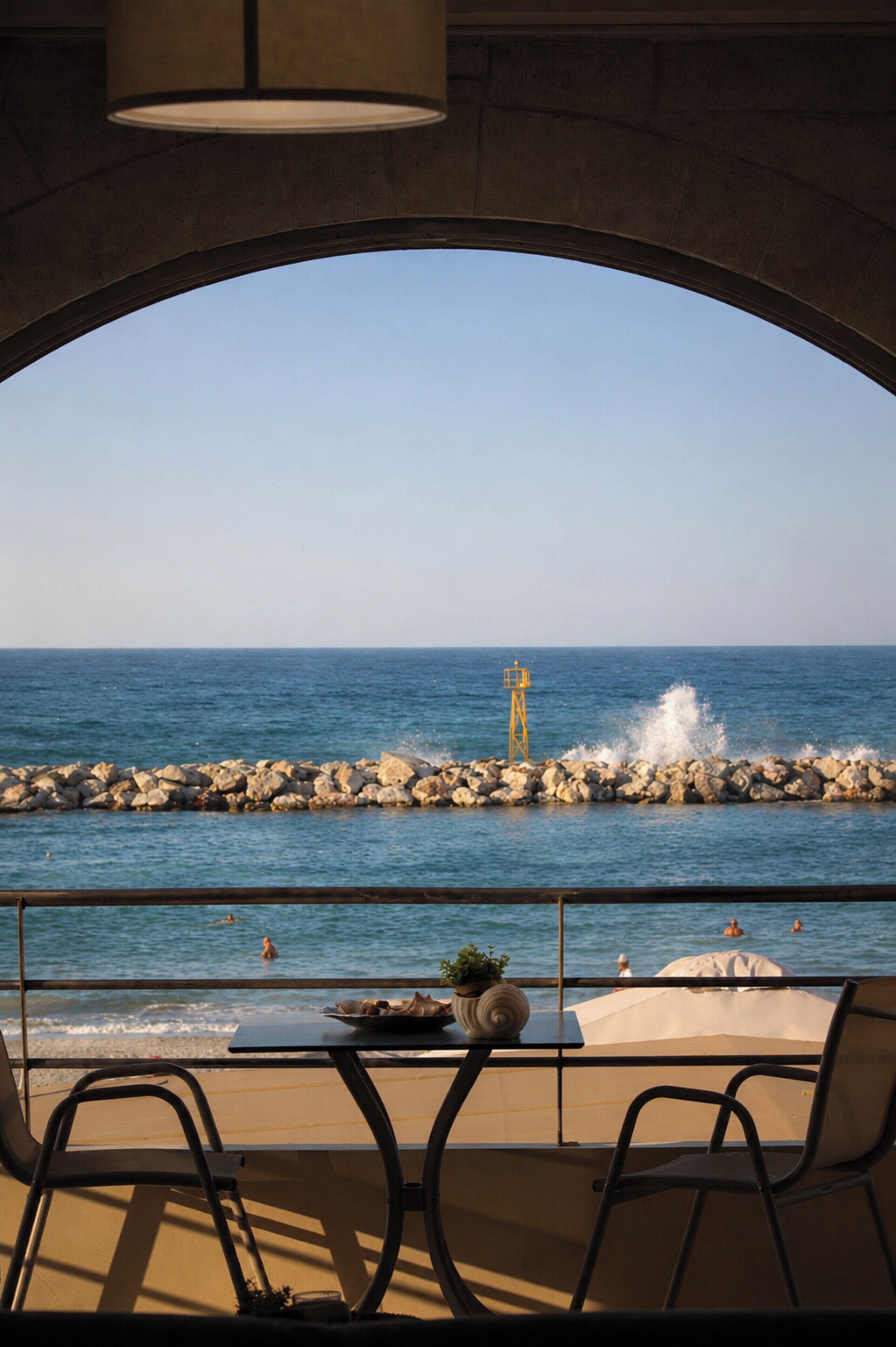 A view from a balcony with a table and chairs, overlooking the sea and a rocky breakwater.