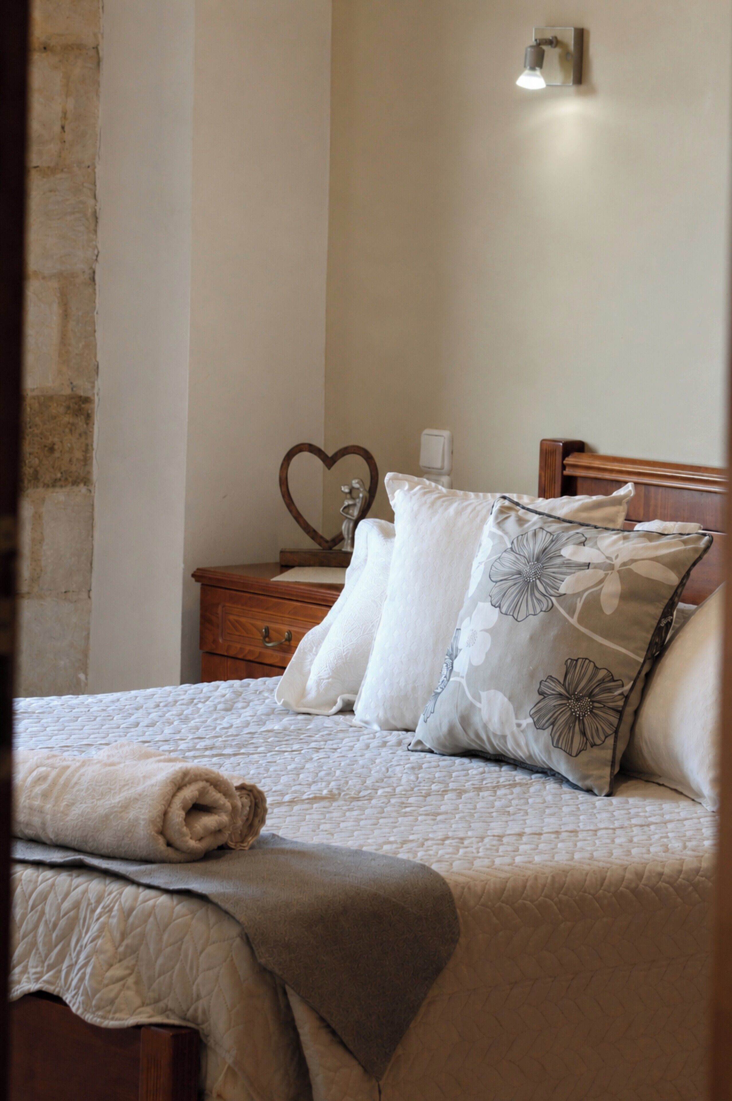 Ein ordentlich bezogenes Bett mit einer gesteppten Decke, ein Nachttisch aus Holz mit einem herzförmigen Spiegel und eine Wandleuchte.