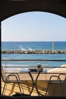 Vista desde un balcón con vista al mar, que incluye una mesa con sillas, una planta en maceta y un rompeolas rocoso en la distancia.