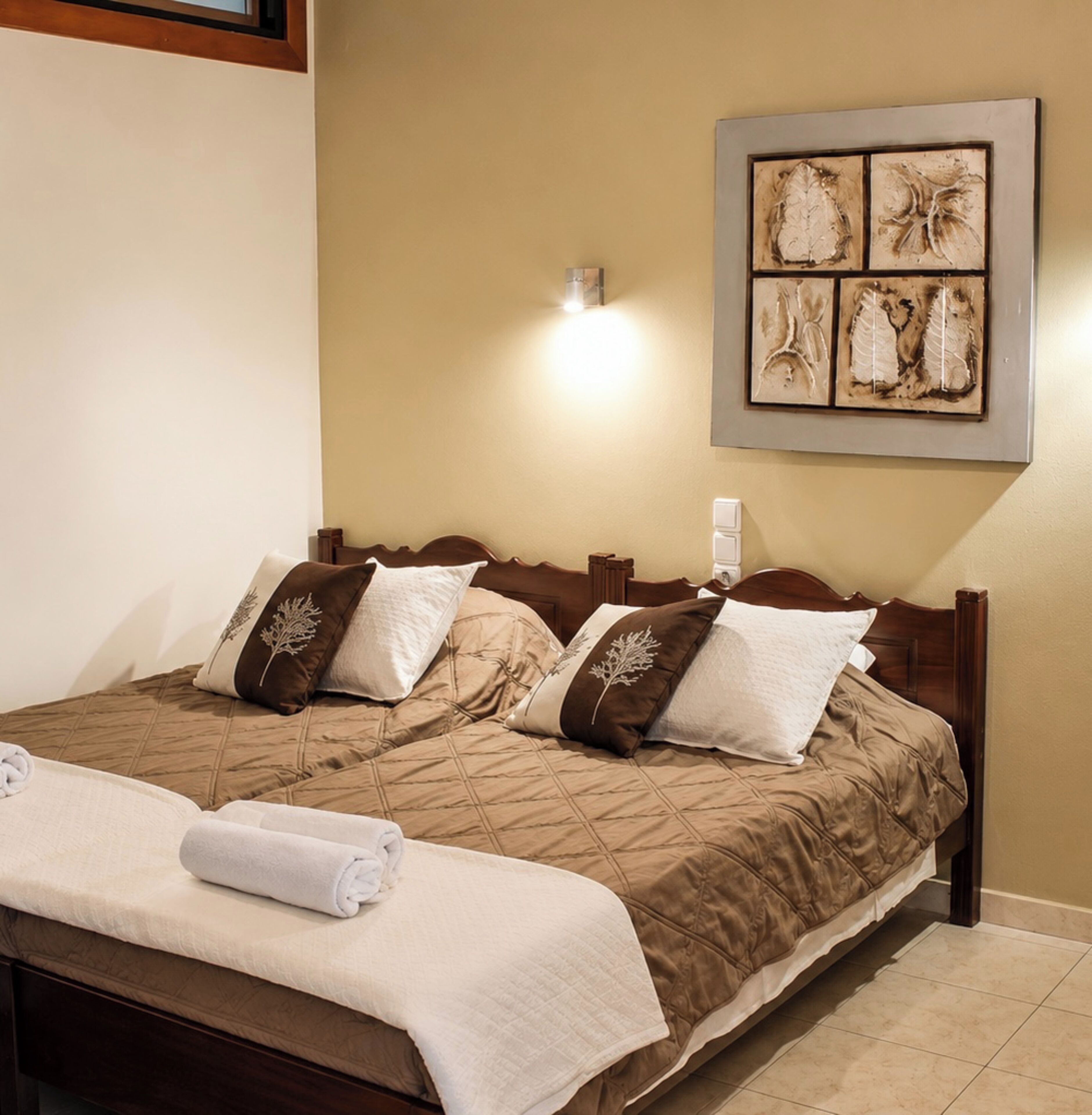 Una cama doble con una colcha marrón acolchada, dos almohadas decorativas, una lámpara de pared y una obra de arte enmarcada sobre la cama.
