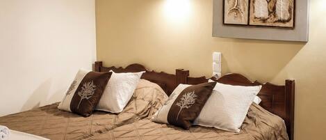 Una cama doble con una colcha marrón acolchada, dos almohadas decorativas, una lámpara de pared y una obra de arte enmarcada sobre la cama.