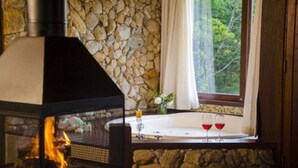 Una chimenea de piedra con fuego encendido, un muro de piedra, una ventana con vistas a la vegetación y una bañera con dos copas de vino tinto.