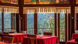 Un restaurante con varias mesas cubiertas por manteles rojos, amplias ventanas con cortinas blancas y vistas a un paisaje boscoso.