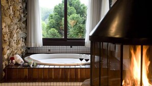 Un baño con pared de piedra, una bañera, un elemento decorativo con fuego y vistas a árboles a través de una ventana.