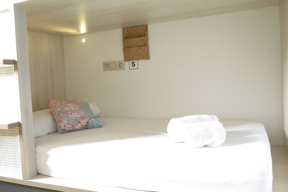 A single-bed room with a wooden headboard, a floral pillow, a white blanket, and a small wooden shelf above the bed.